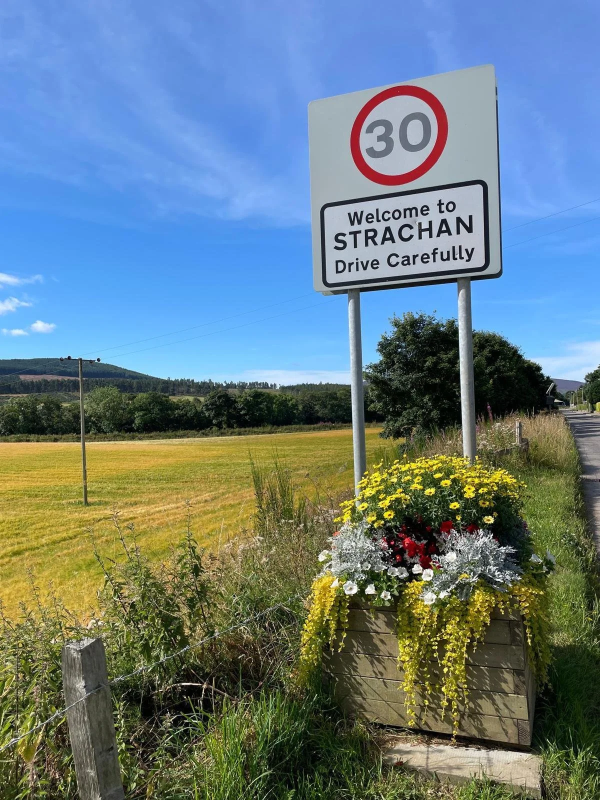 Road sign welcoming to Strachan Village with a speed limit of 30 mph, next to a flower box with yellow, red, white, and grey flowers on a grassy roadside under a blue sky. Flower box provided by Strachan Flowers group. Feughside Community Association