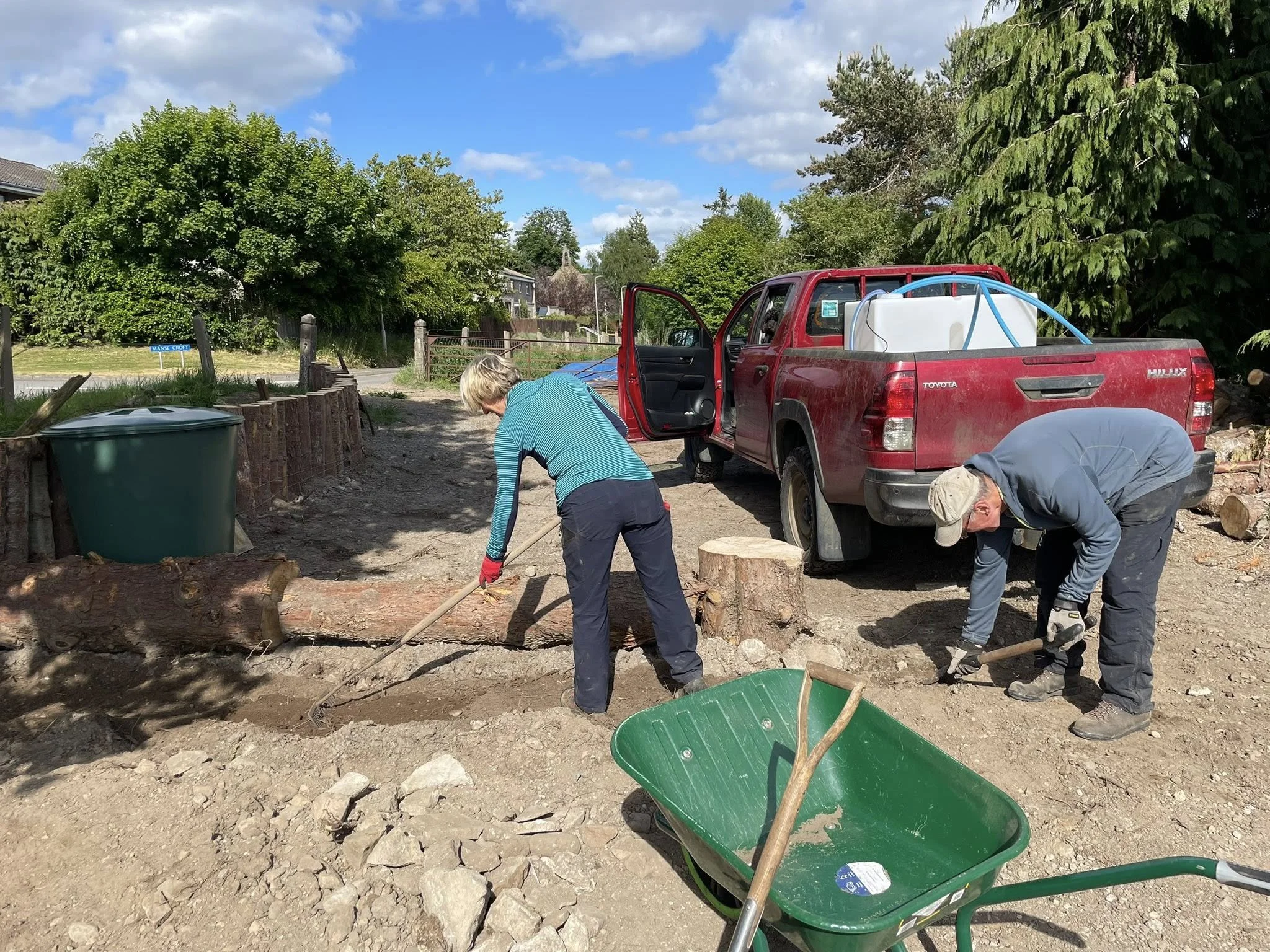 Feughside/Strachan Community Garden. A woman and man, digging in the dirt near a red pickup truck with a water tank in the truck bed. There is a green wheelbarrow and a large trash bin nearby, and a tree-lined area in the background.