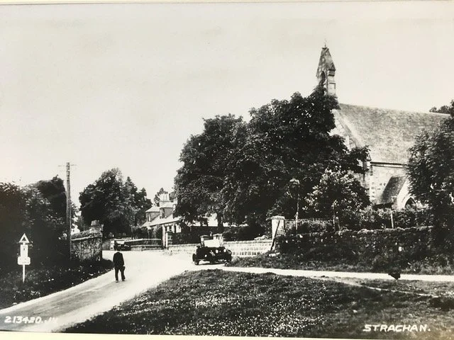 A black and white photo of a rural or small-town scene with a church, trees, and a few buildings. A person is walking on a dirt or gravel road, and a vintage car is parked nearby.