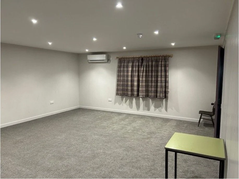 Empty Strachan Hall small Hall with grey carpet, white walls, a window with plaid curtains, an air conditioner, and a few chairs and a table.