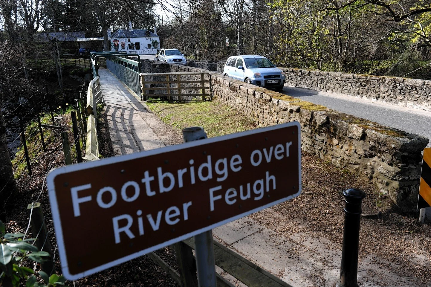 Footbridge over River Feugh with cars parked on the road, stone walls along the bridge, a pedestrian pathway, and a sign indicating the footbridge over River Feugh.