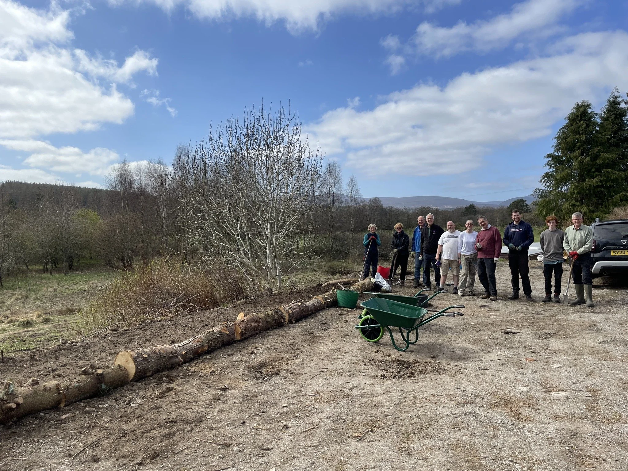 Feughside/Strachan Community Garden. Group of people standing outdoors on a dirt area with trees and hills in the background, surrounded by tools and wheelbarrows, apparently working on a landscaping or tree planting project.