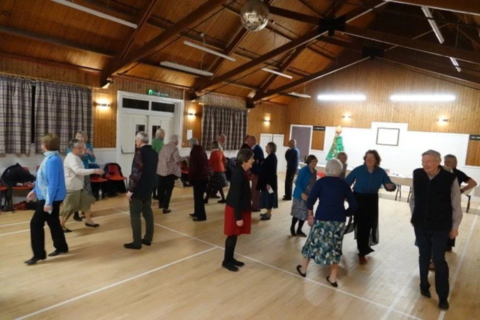 Older adults dancing and socializing in a community hall with wooden walls and ceiling, and a Christmas tree in the background.