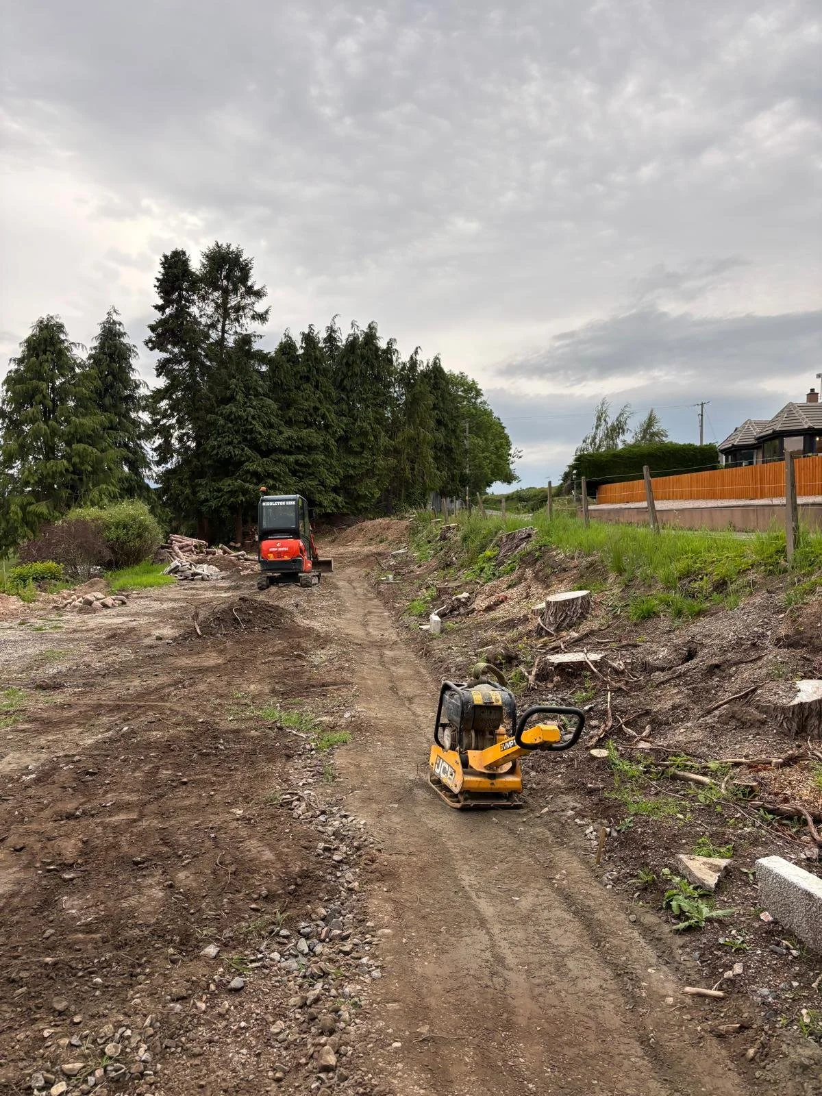 Feughside/Strachan Community Garden. Construction site with dirt road, a small yellow plate compactor in the foreground, a small excavator further back, and trees with green leaves on the left side of the image under a cloudy sky.