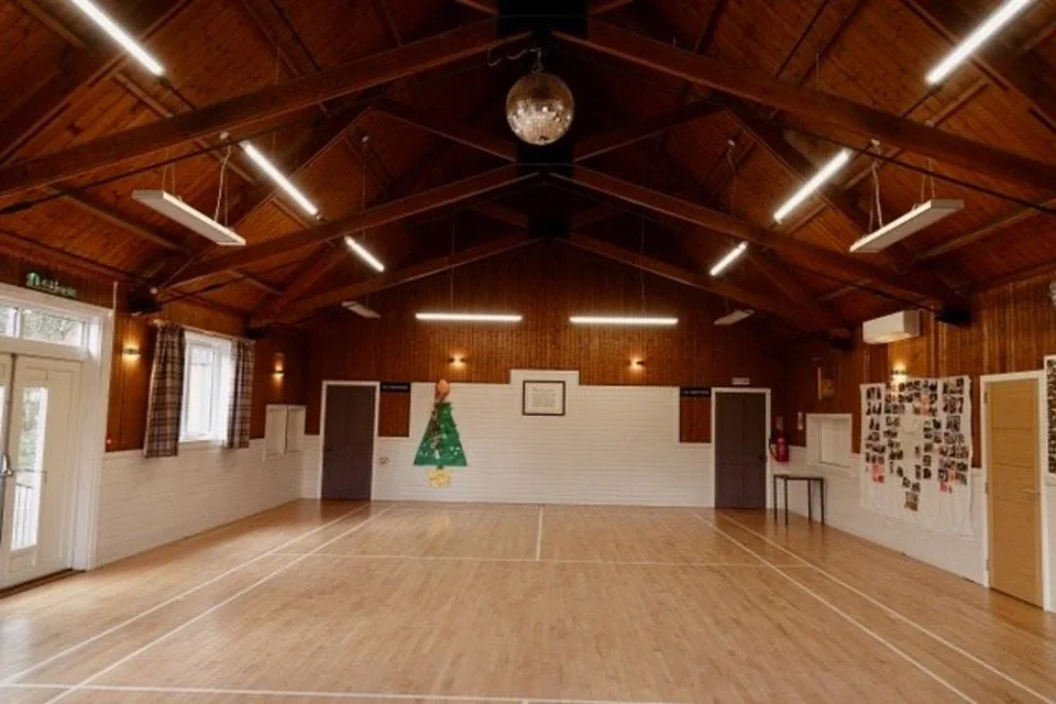 Empty Strachan Main Hall (big Hall) with wooden floor and wood-paneled walls and ceiling, decorated with a Christmas tree cutout, photographs, and a framed certificate