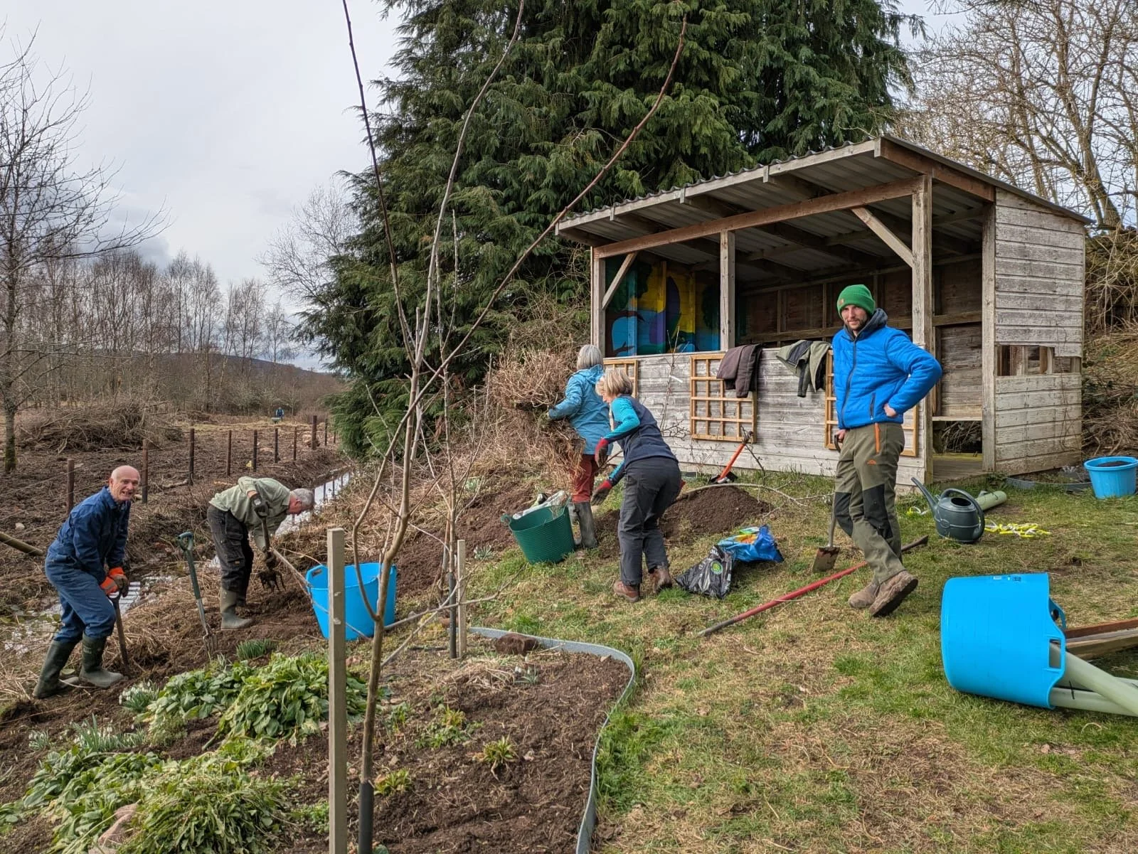 Group of six people working together gardening or landscaping at Feughside community garden in Strachan, near a wooden shed, with hand tools and watering cans, on a cloudy day.