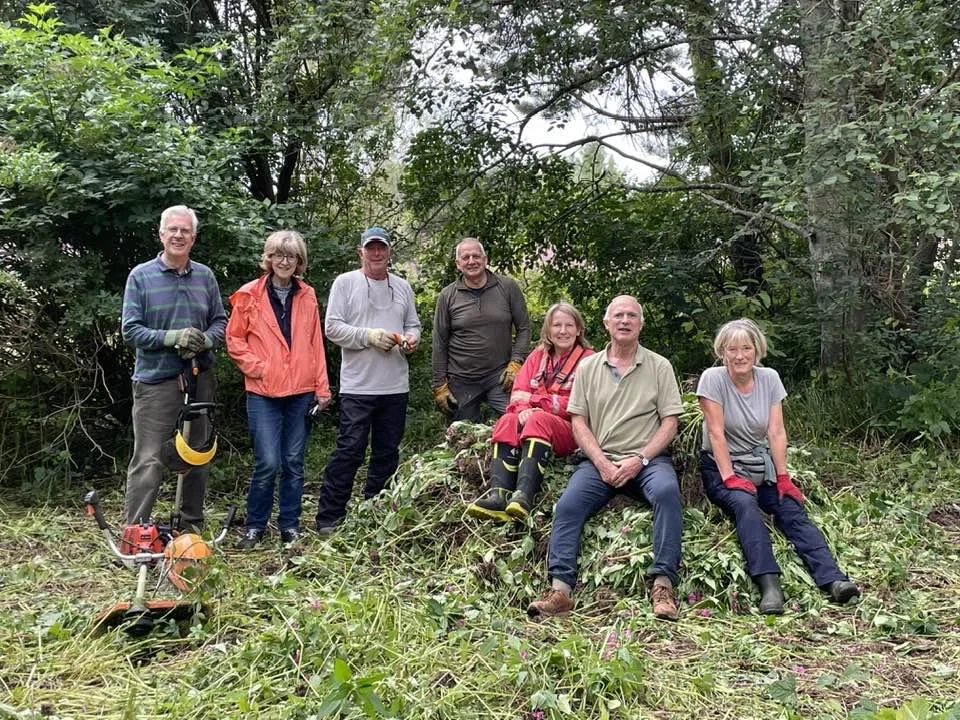 Feughside/Strachan Community Garden. Group of seven people outdoors in a forest, some sitting on a small mound, others standing, surrounded by greenery, wearing work gloves and casual clothes, with gardening tools visible.