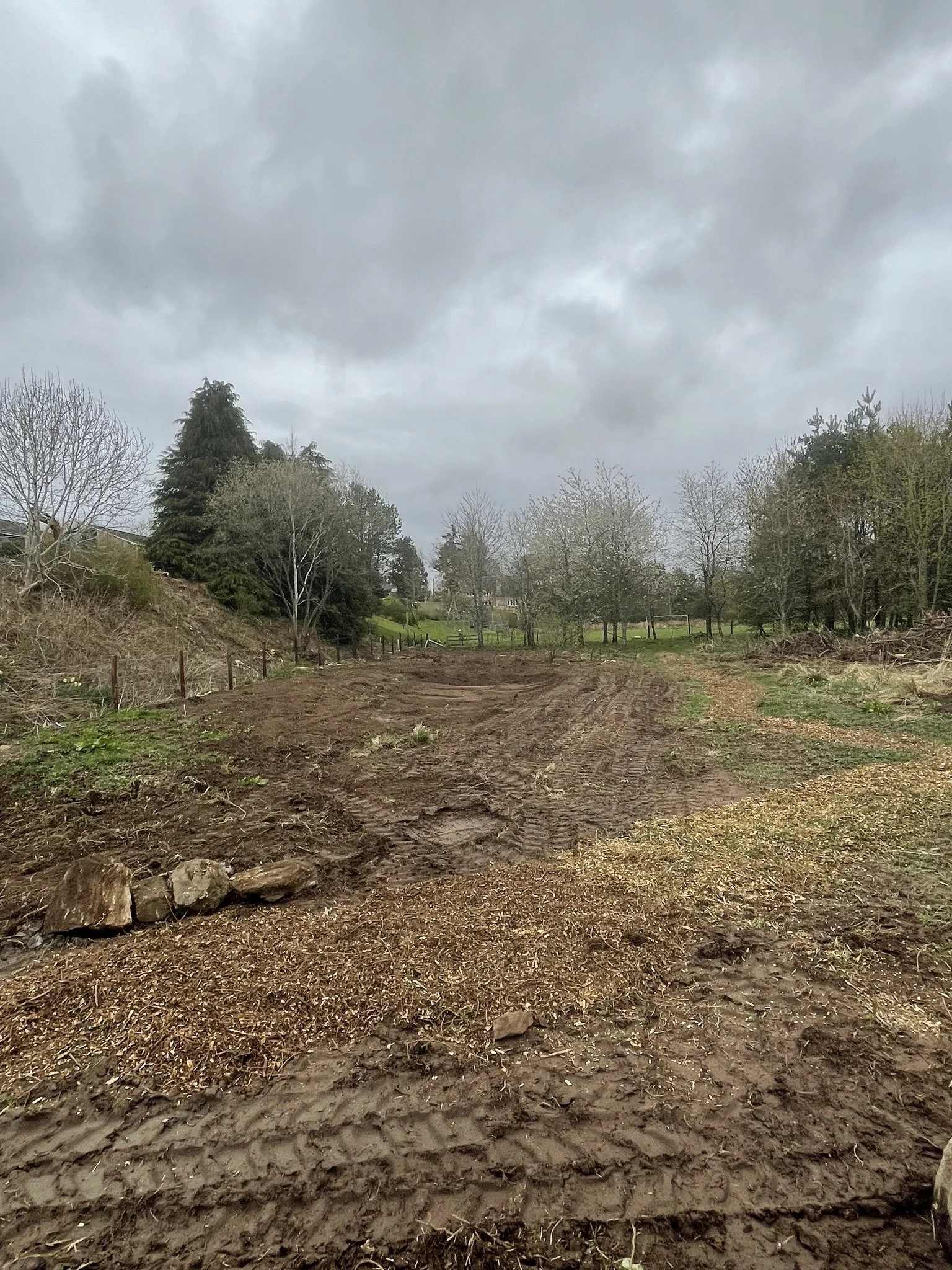 Feughside/Strachan Community Garden. A dirt path or field with tire tracks, bordered by rocks and sparse grass, leading towards a green area with trees under a cloudy sky.