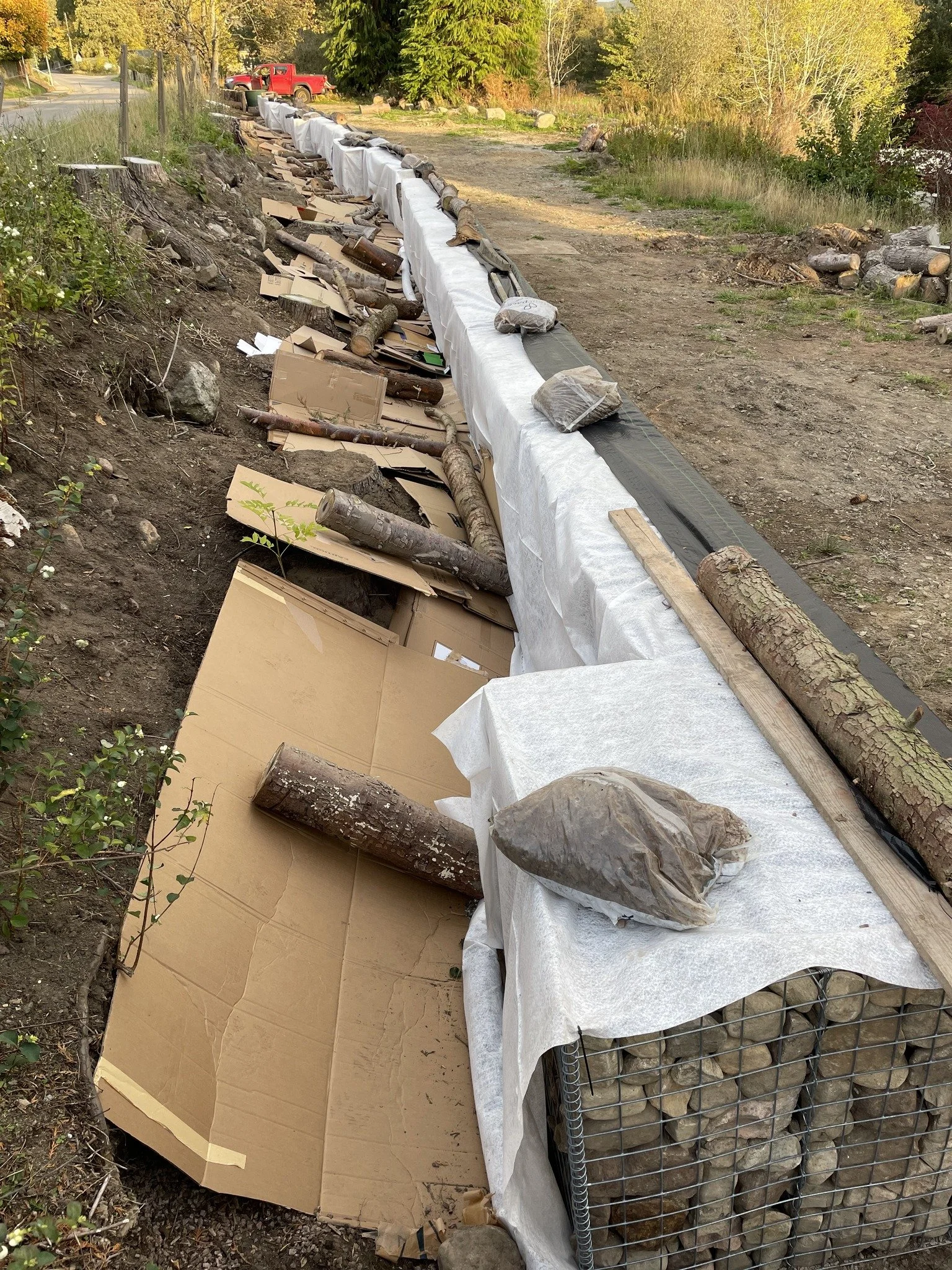 Feughside/Strachan Community garden. A long outdoor construction project with logs, rocks, and cardboard on the ground, and a red truck in the background.