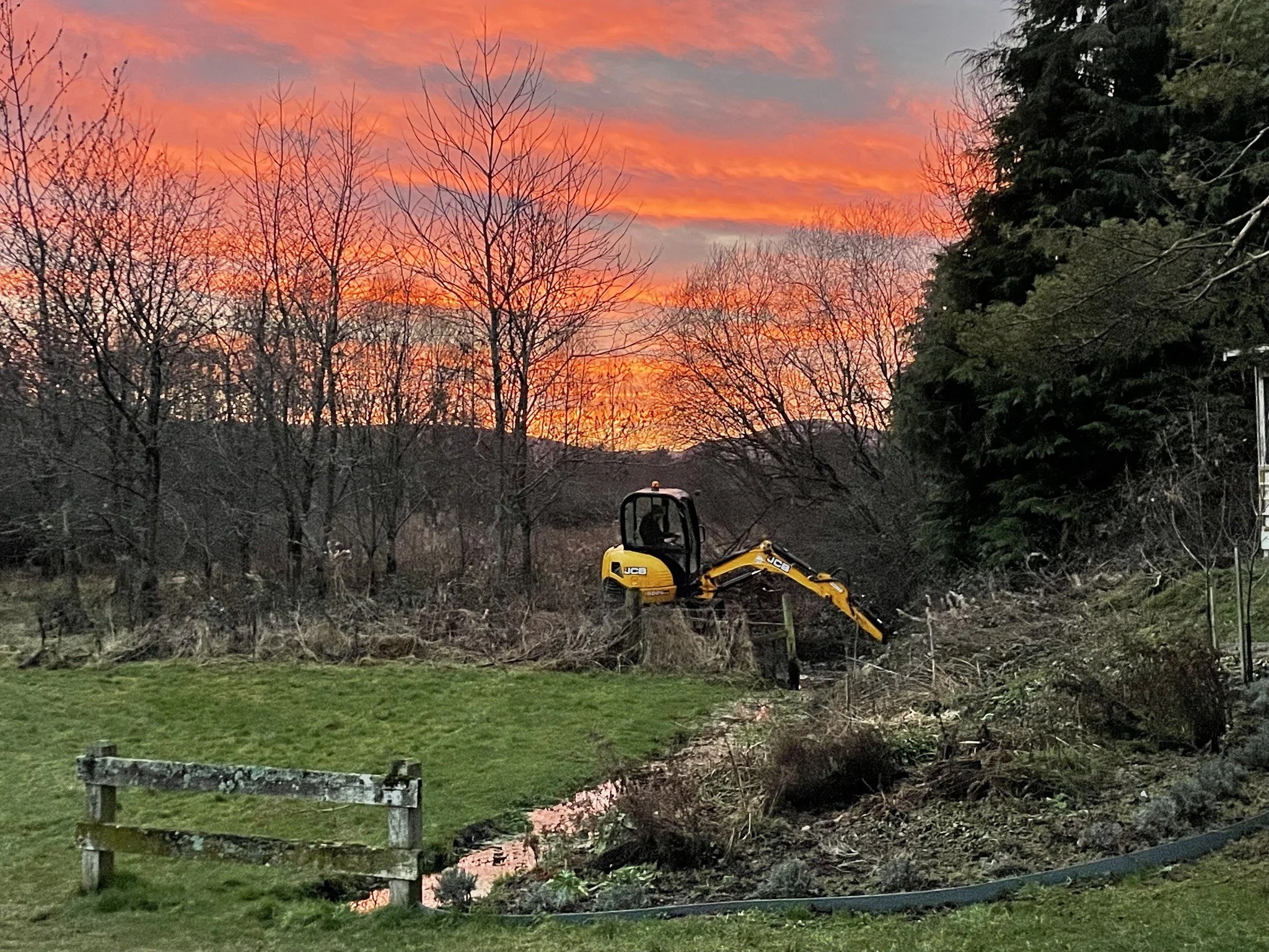 Feughside/Strachan Community Garden. A small yellow excavator working in a garden area during sunset, with a colorful orange and pink sky and bare trees in the background.