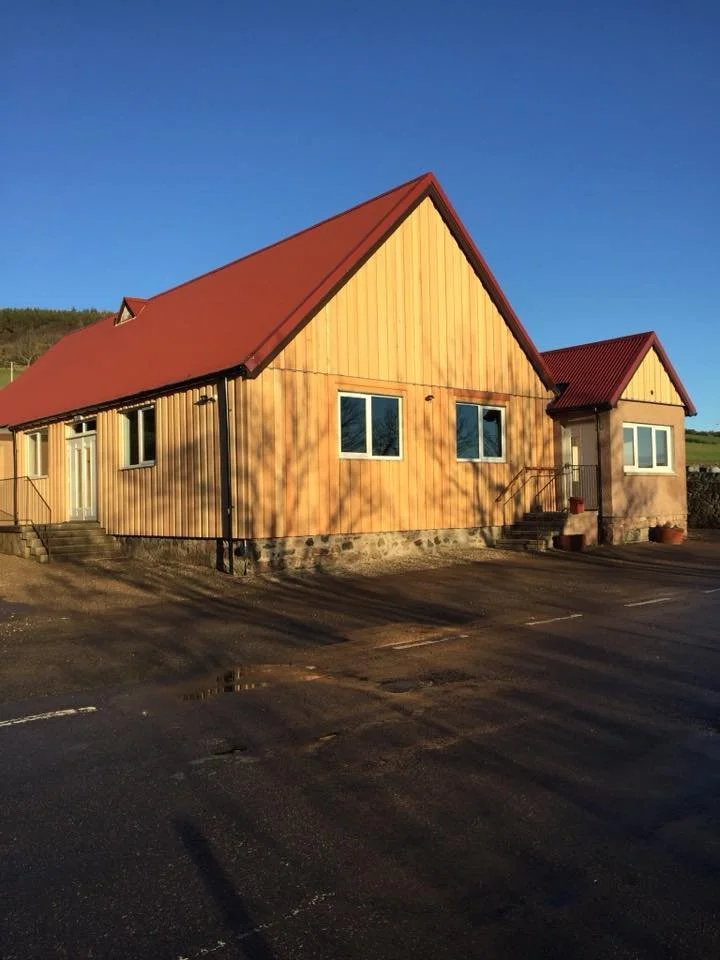 A wooden building with a red metal roof and several windows, situated on a paved lot under a clear blue sky.
