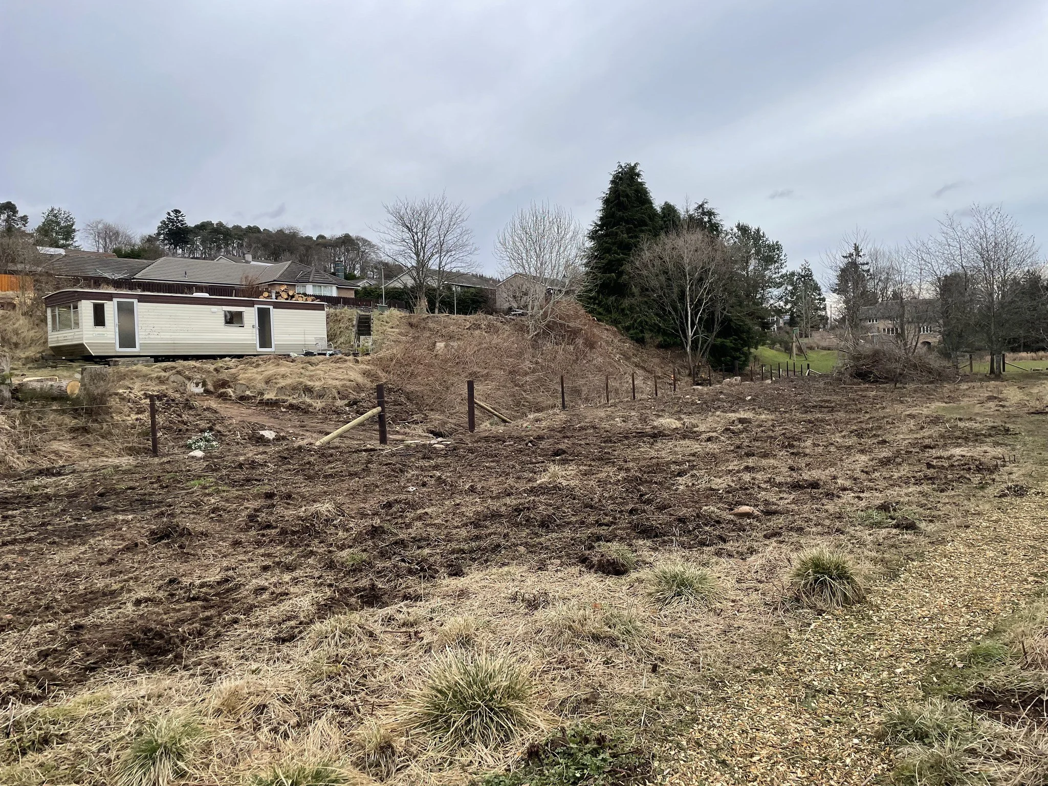 Feughside/Strachan Community Garden. A cleared plot of land with patches of grass and dirt, some small shrubs, and a few trees in the background, with a white trailer on the left side, under an overcast sky.