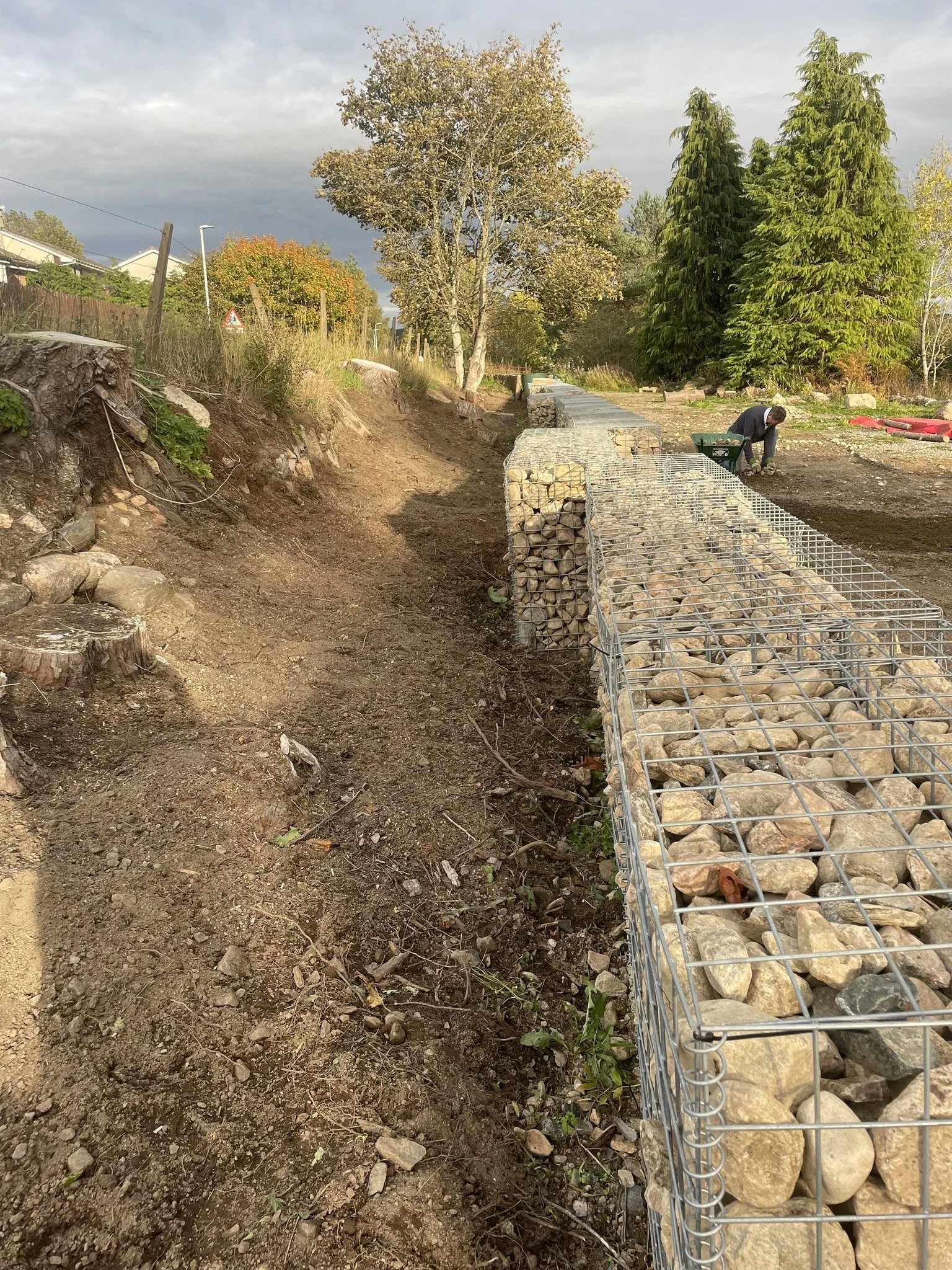 Feughside/Strachan Community Garden. A construction site showing a path lined with gabion baskets filled with rocks, with a worker in the background, trees, and a cloudy sky.