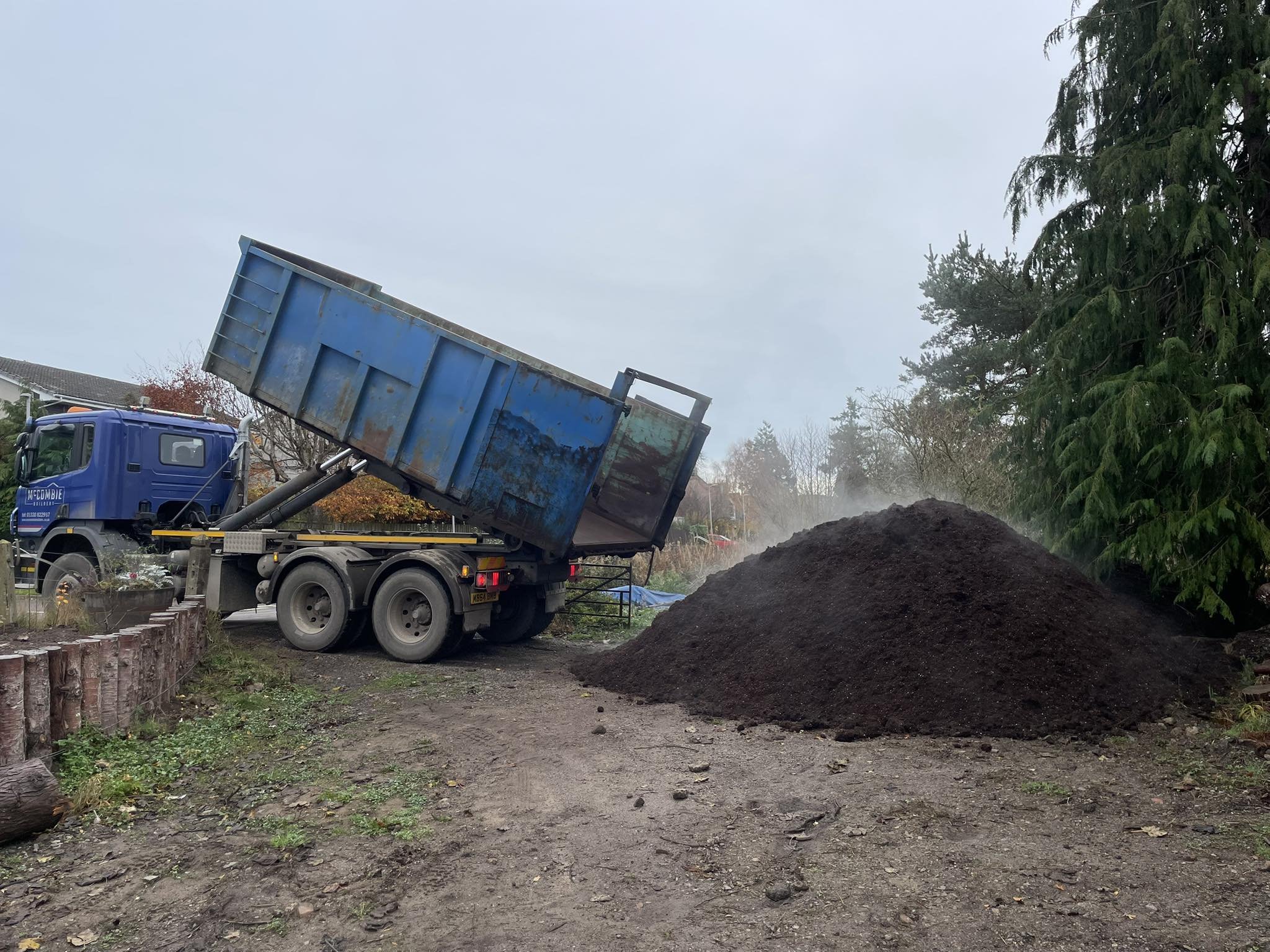 Feughside/Strachan Community Garden. A blue dump truck unloading a pile of dark soil or compost onto a dirt ground outdoors, with trees and houses in the background.