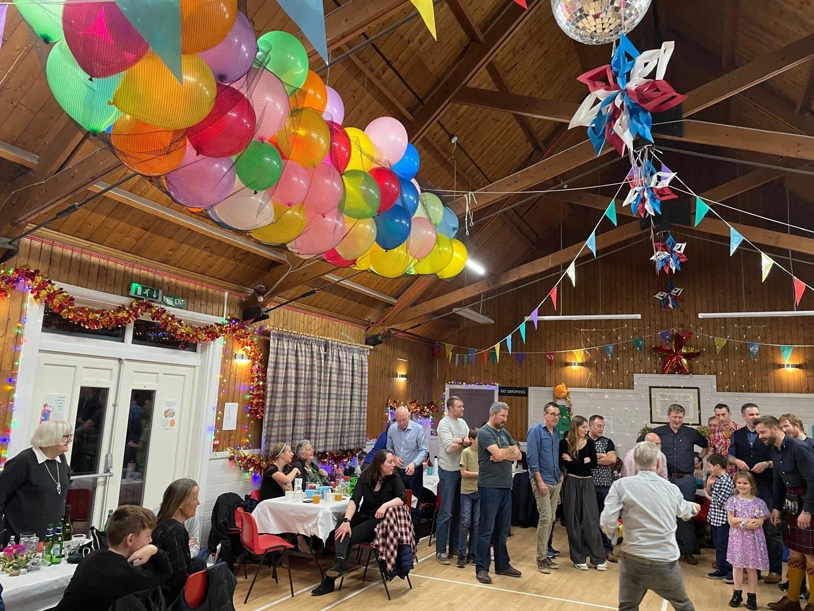 A lively celebration at Strachan Hall with festive decorations, balloons, and colorful bunting hanging from the ceiling. Guests are seated at tables and standing, engaged in conversation and enjoying the event.