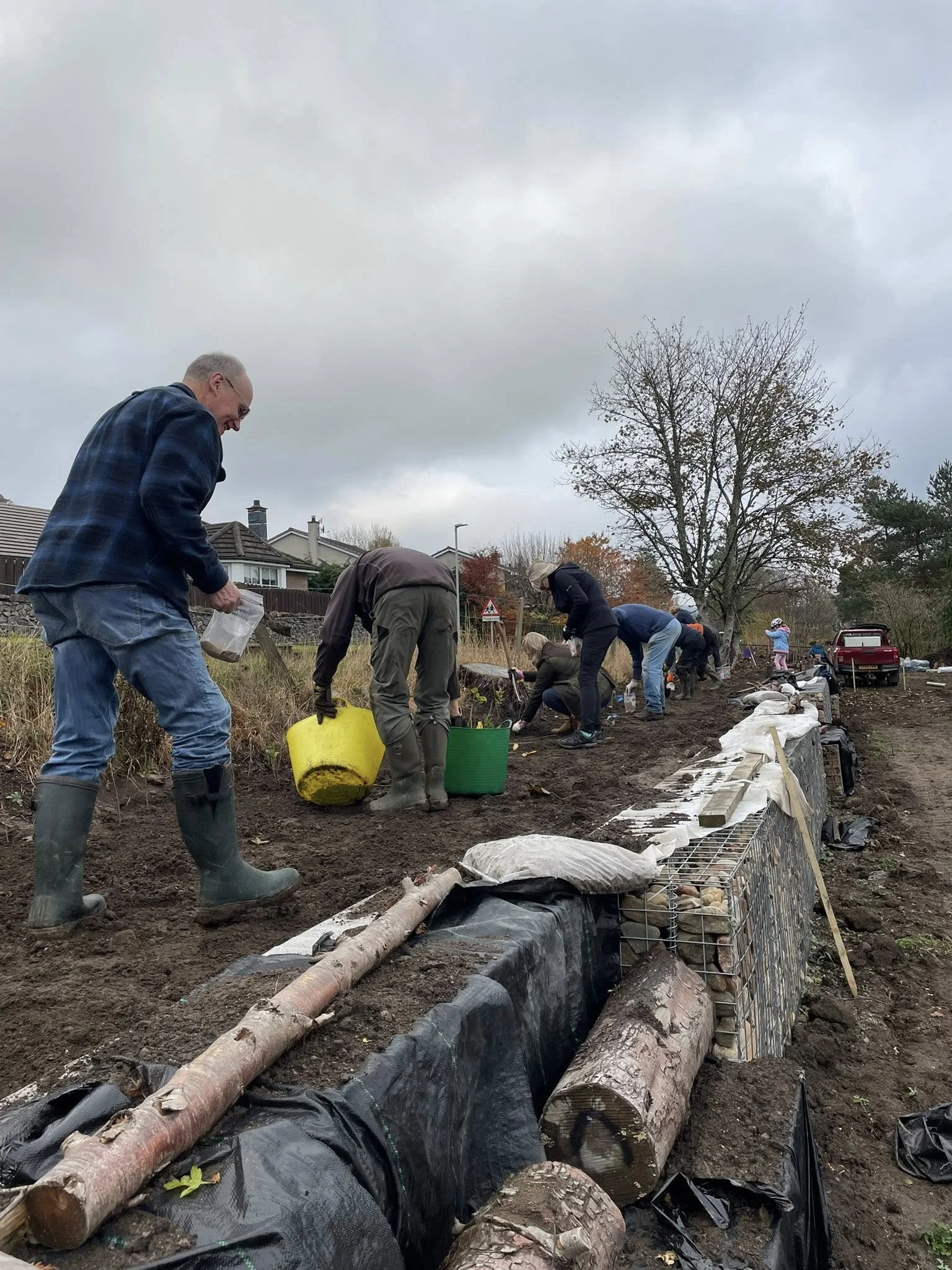 Feughside/Strachan Community garden. People planting trees or working on a garden along a soil embankment on a cloudy day.