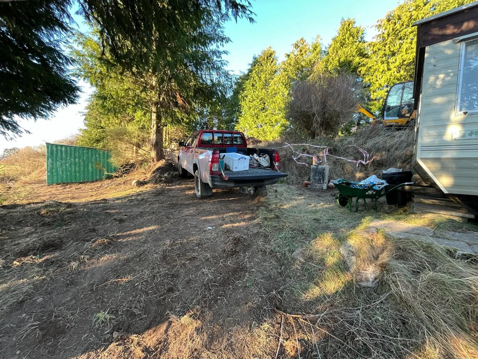 Feughside/Strachan Community Garden. A parked red pickup truck carrying storage containers, on rocky ground under trees, with a cloudy sky, next to a trailer, a green wheelbarrow, and construction equipment.