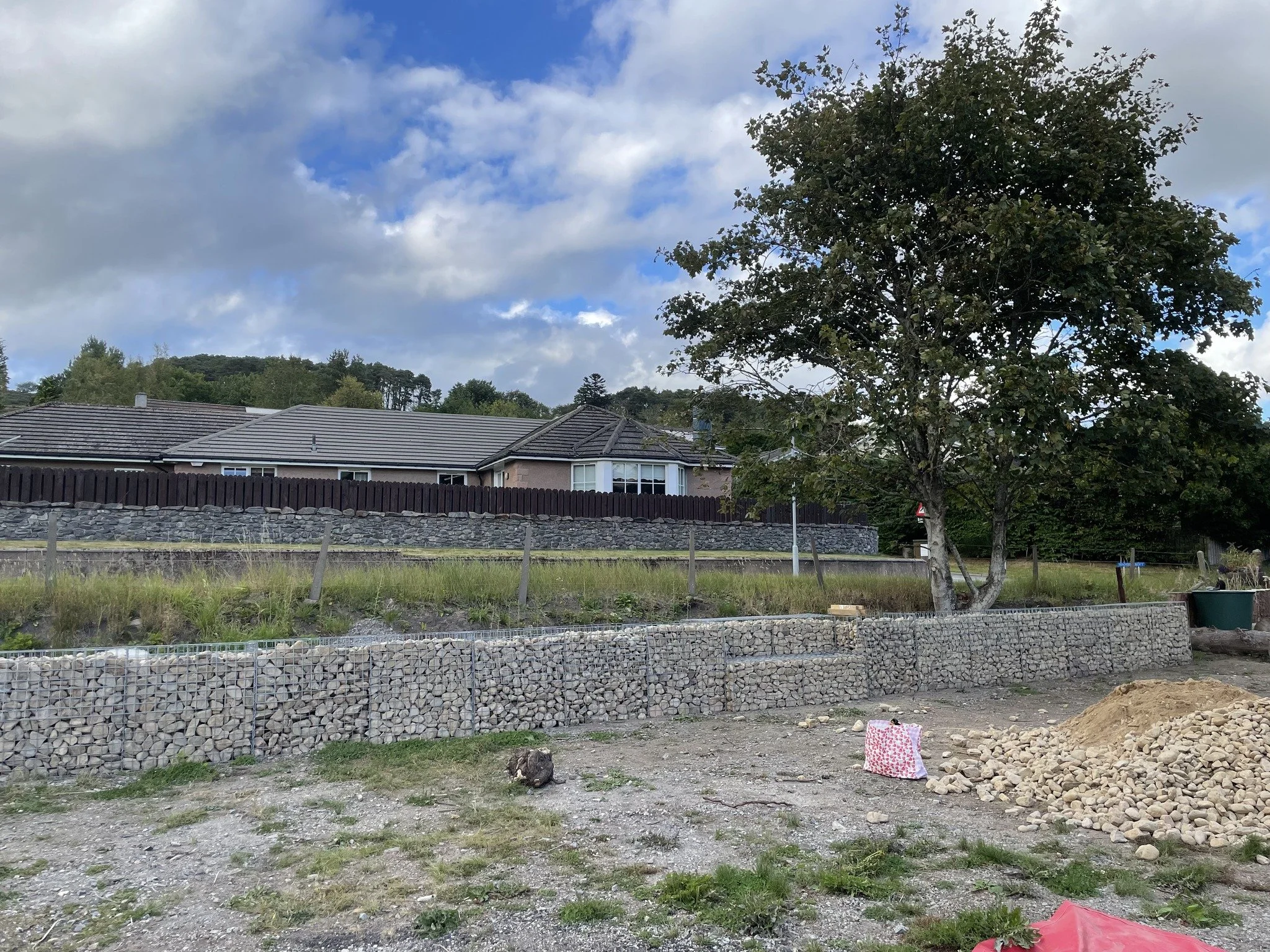 Feughside/Strachan Community garden. A backyard with a large tree, tall wire gabion walls filled with rocks, and construction materials including rocks, a bag, and gravel on the ground.
