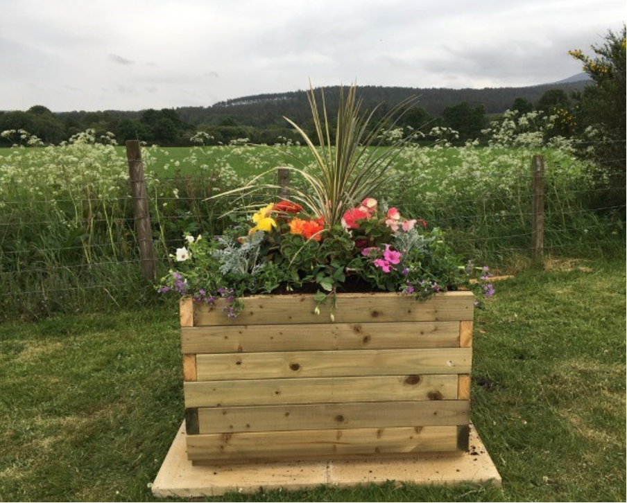 A wooden planter box filled with colourfu flowers, including pink and white blooms, tall grasses, and purple flowers, outdoors in a grassy yard in Strachan, with a fence and a field with trees and hills in the background. By Strachan Flower Group.