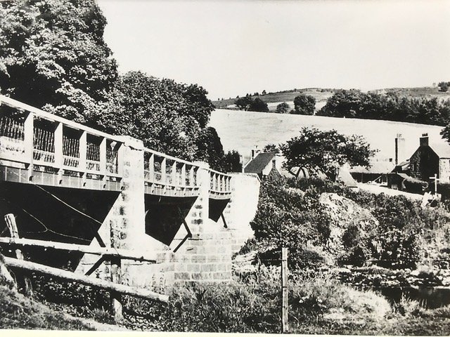 A black and white photograph of a rural landscape featuring a bridge, trees, fields, and a few houses with chimneys in the distance.