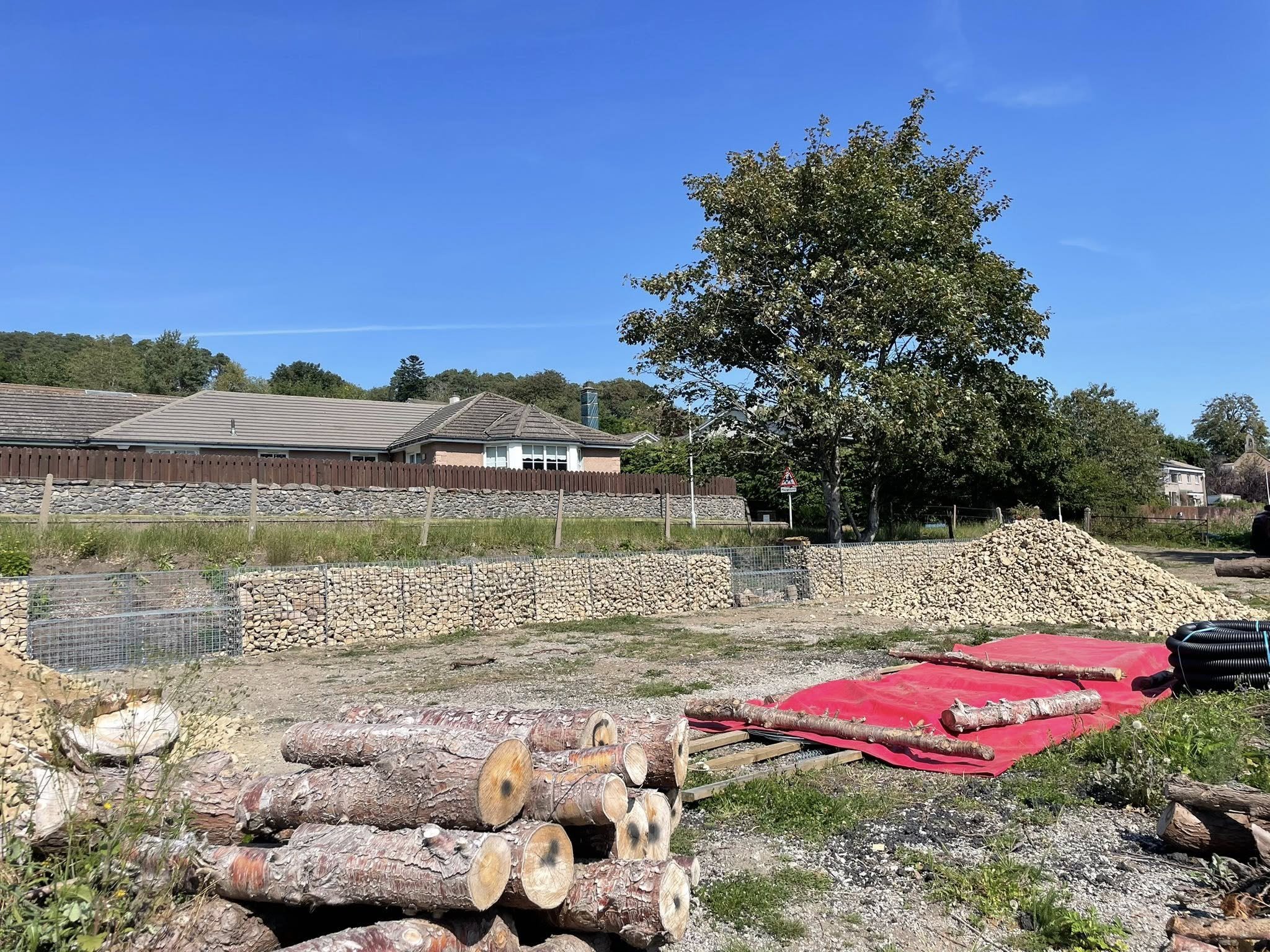 Feughside/Strachan Community Garden. A construction site with a pile of logs, a red tarp, and a large pile of rocks. There are two gabion wall structures filled with rocks, a large tree, houses in the background, and a clear blue sky.