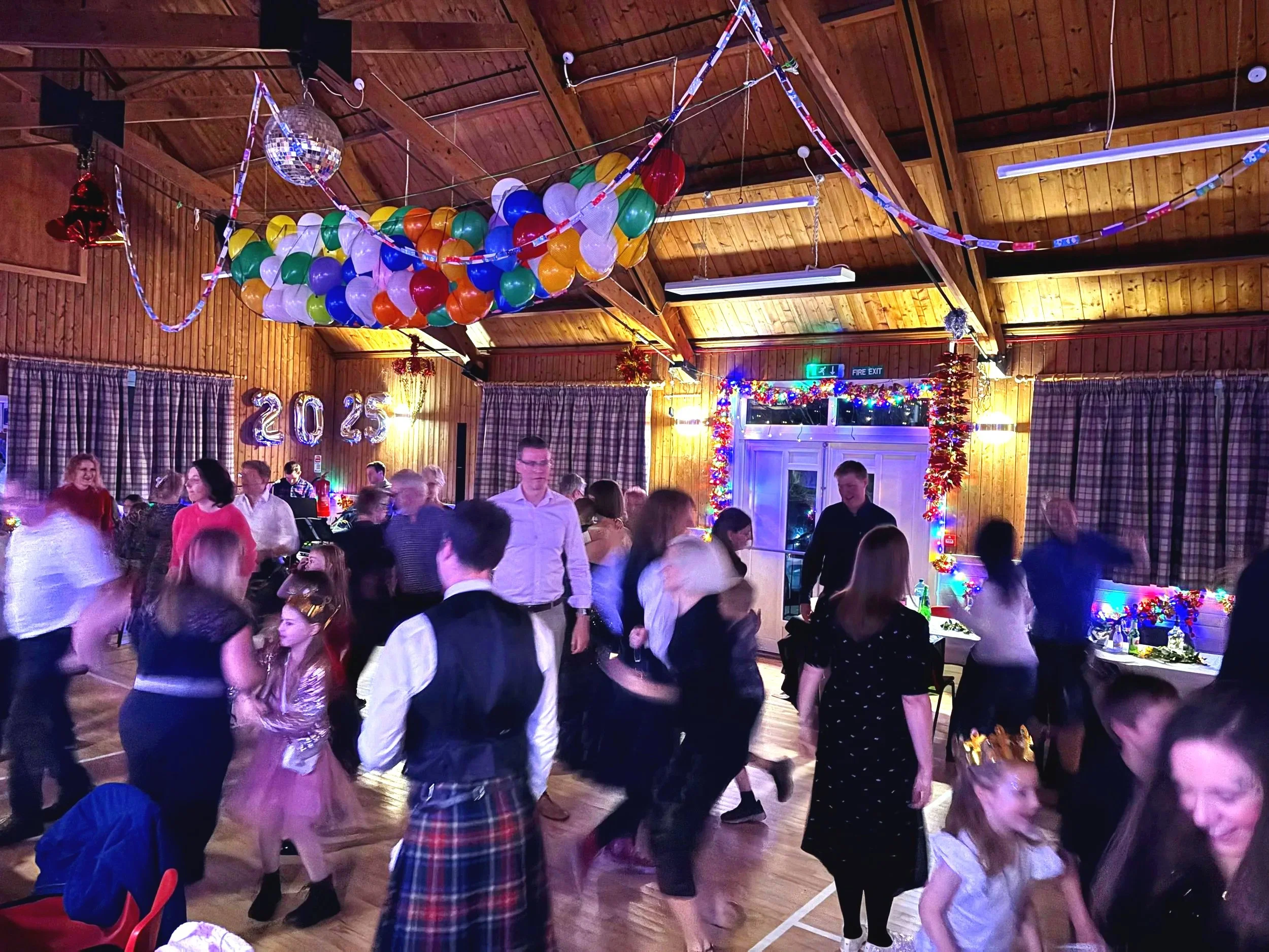 People dancing and socializing in a wooden hall decorated for a celebration with balloons, colorful lights, and '2023' balloons on the wall.