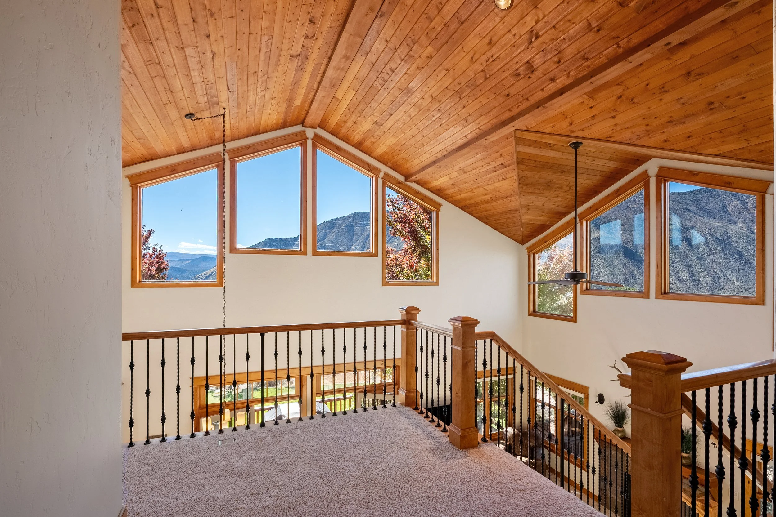 Upper level of a house with wooden ceiling and large windows showing mountains and trees outside. Carpeted floor and black metal railing around the open space overlooking the lower level.