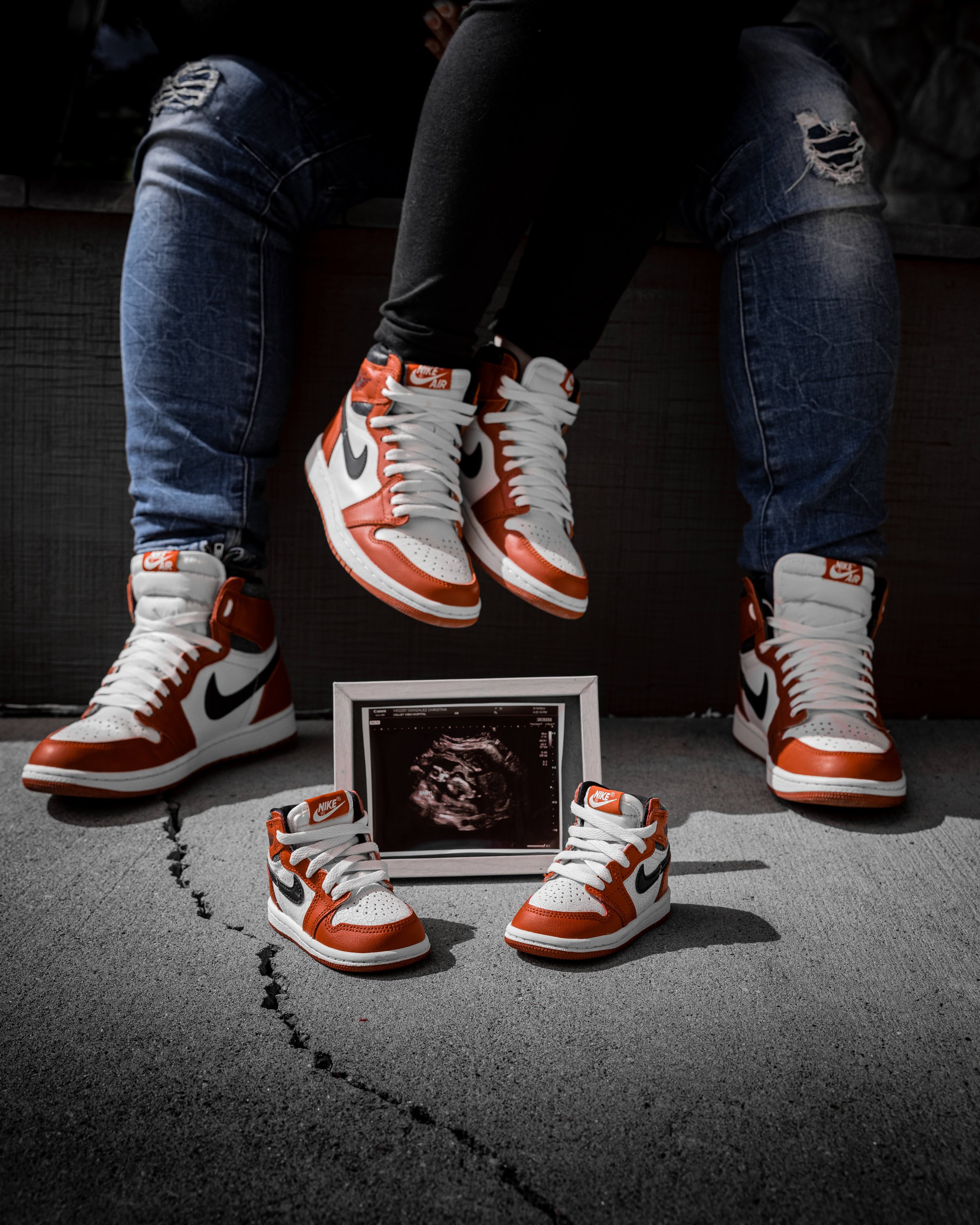 Two people wearing matching red, white, and black Nike sneakers, sitting with their legs extended. In front of them is a framed ultrasound image of a fetus, with a pair of tiny matching Nike shoes placed below the ultrasound.