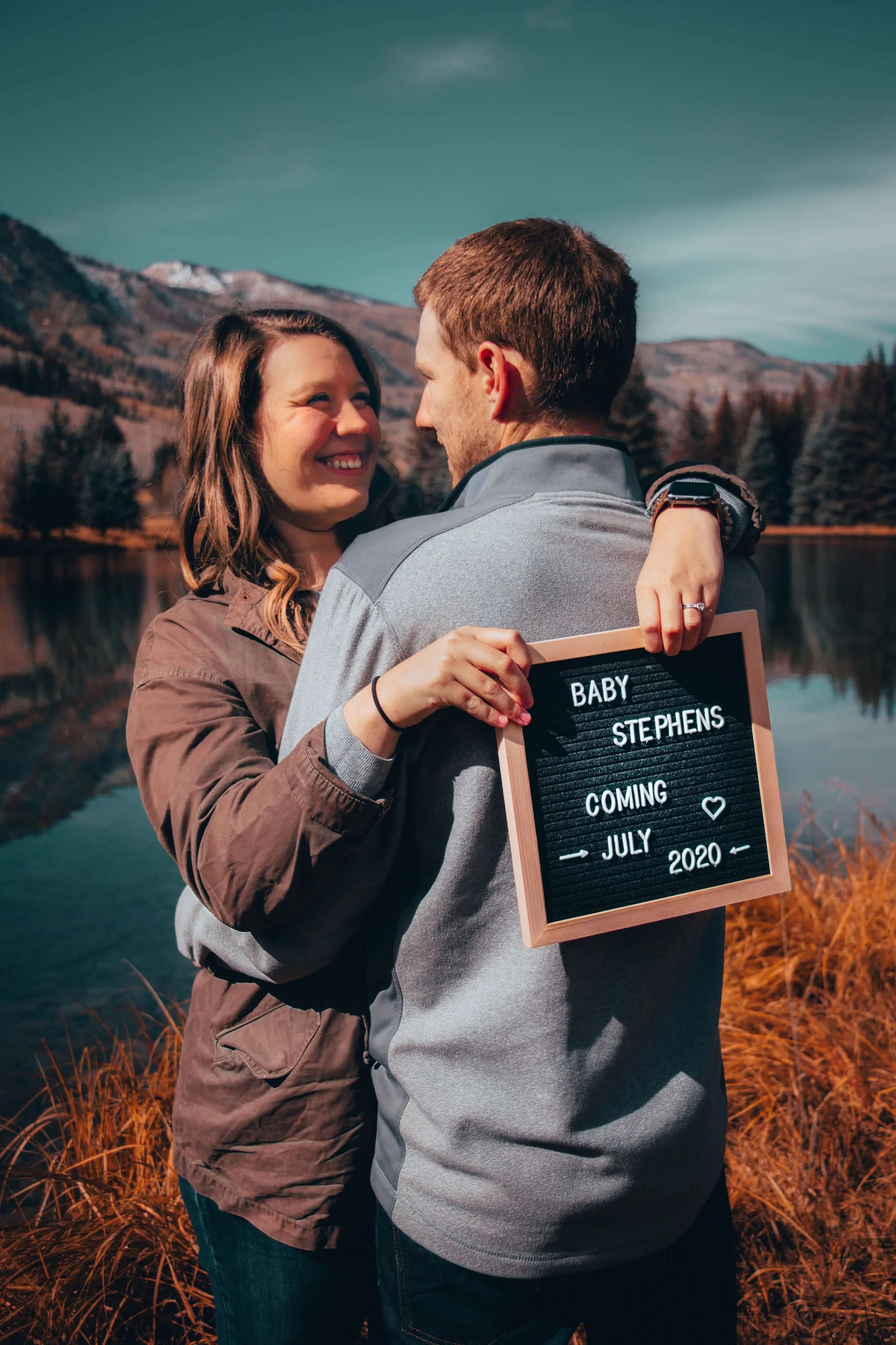 A couple hugging outdoors near a lake with mountains in the background, holding a sign that says "Baby Stephens Coming July 2020".