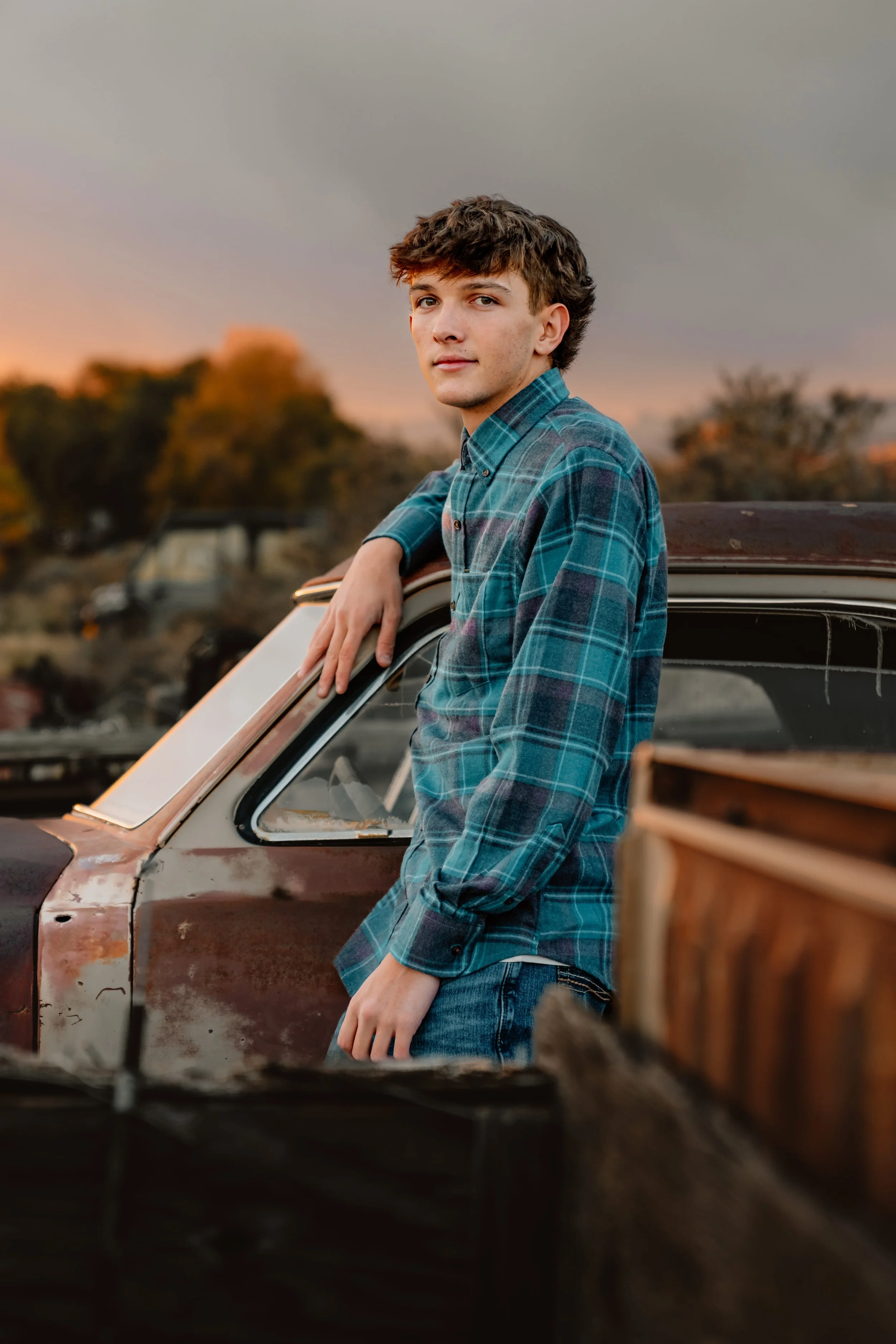 Young man in plaid shirt leaning on an old rusted car outdoors during sunset.