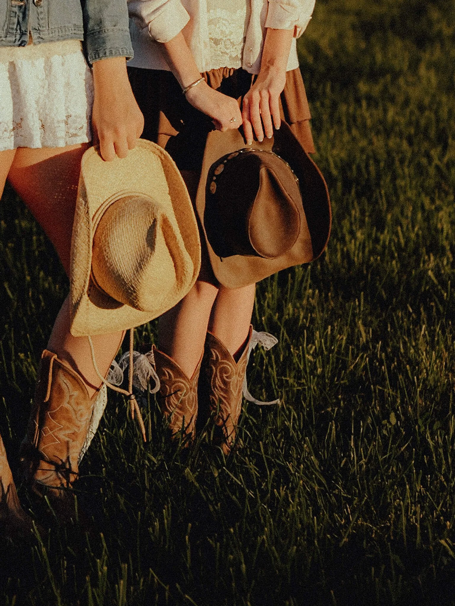 Two people sitting on a grassy field, holding hats and small purses, wearing cowboy boots.