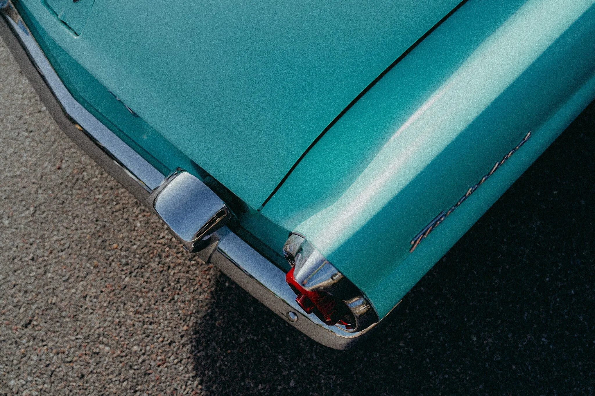 Close-up of the front corner of a vintage teal car, showing the chrome bumper, headlight, and part of the hood with a classic design.