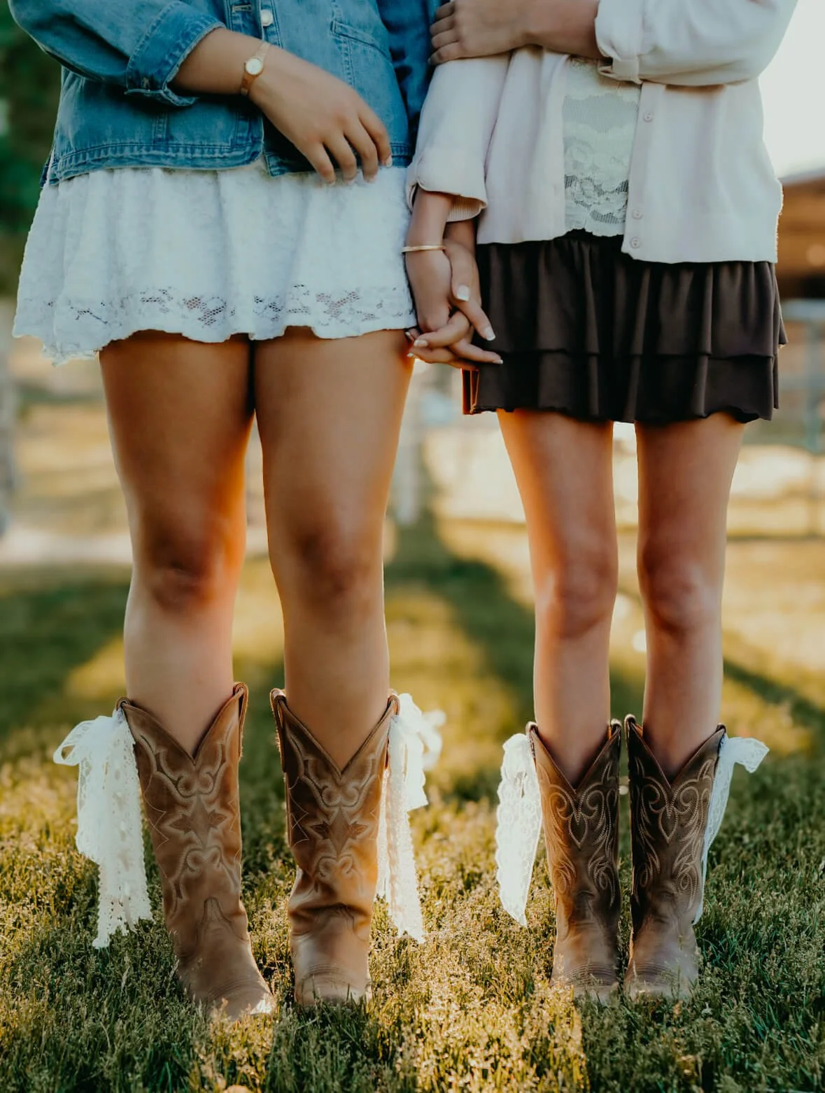 Two women wearing cowboy boots and skirts holding hands outdoors, in a field during sunset.