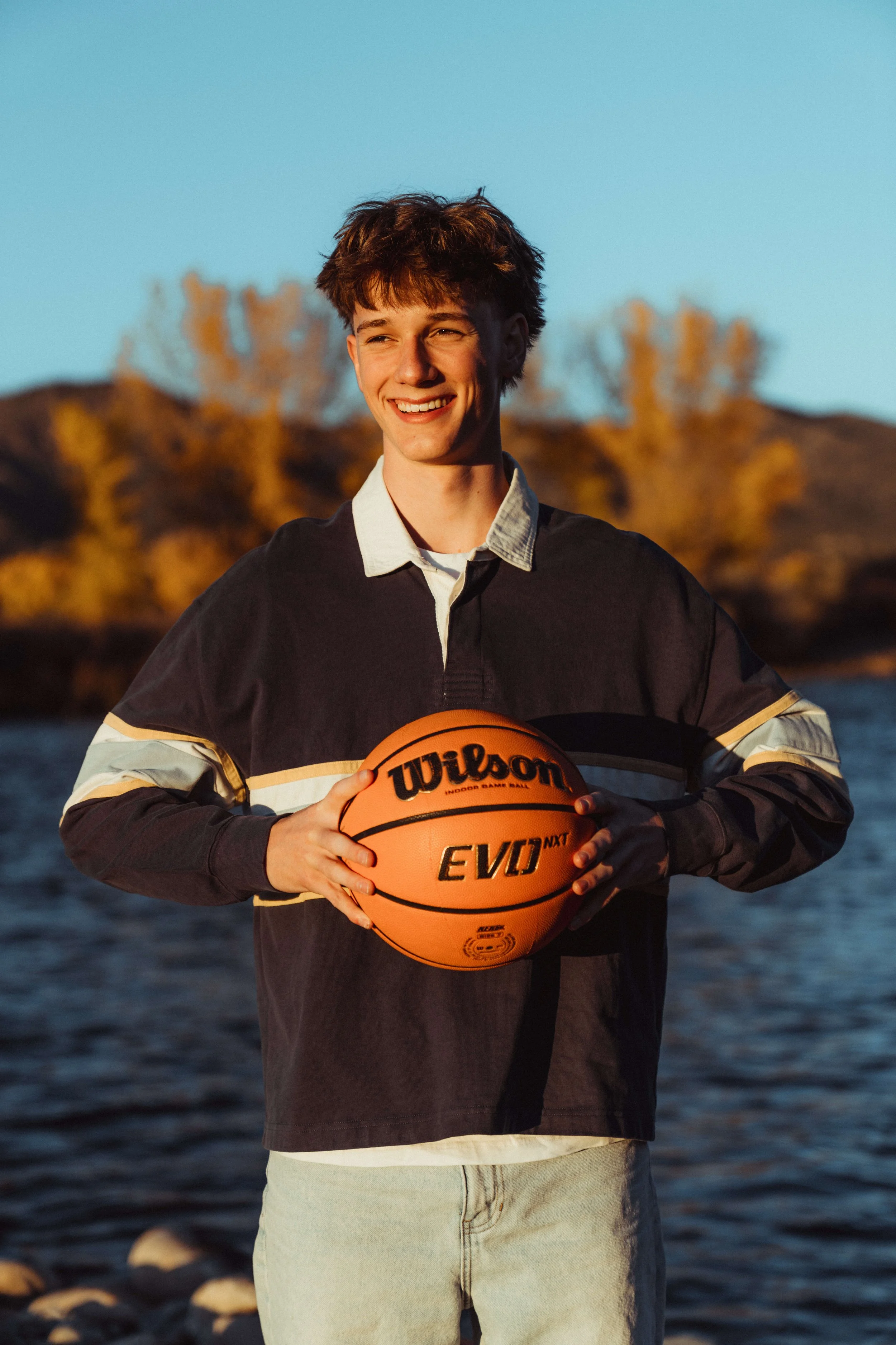 Young man standing outdoors near water, holding a Wilson volleyball, smiling, during sunset.
