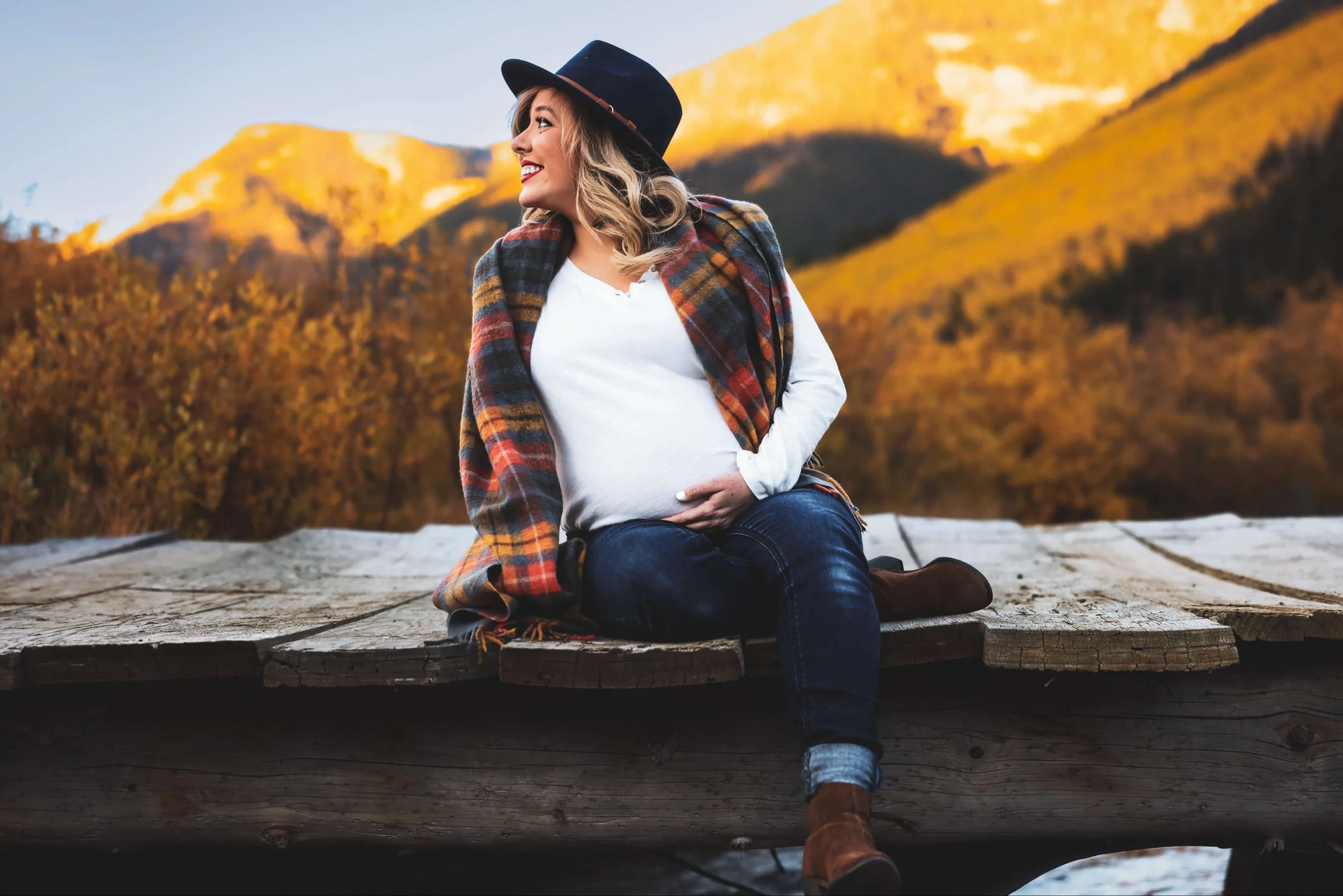 Pregnant woman wearing a hat, sitting on a wooden dock, smiling and looking to the side, with autumn mountains in the background.