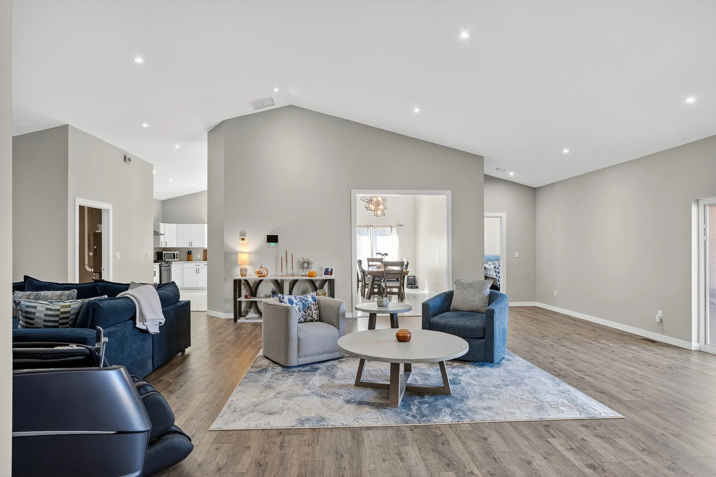 Spacious living room with wooden flooring, light grey walls, and white ceiling with recessed lighting. There are two grey armchairs, a blue sofa, and a large area rug. The room has a minimalistic decor with a small side table, a console table against