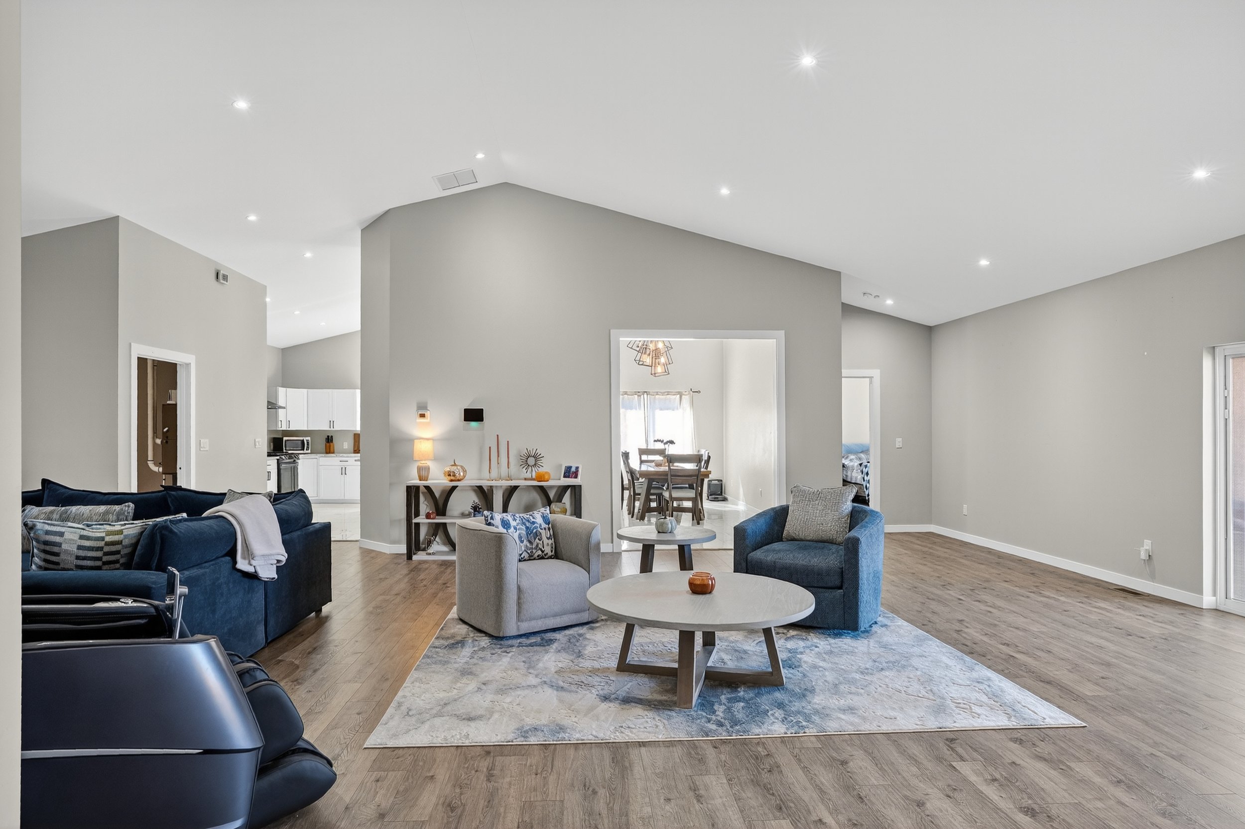 Spacious living room with wooden flooring, light grey walls, and white ceiling with recessed lighting. There are two grey armchairs, a blue sofa, and a large area rug. The room has a minimalistic decor with a small side table, a console table against