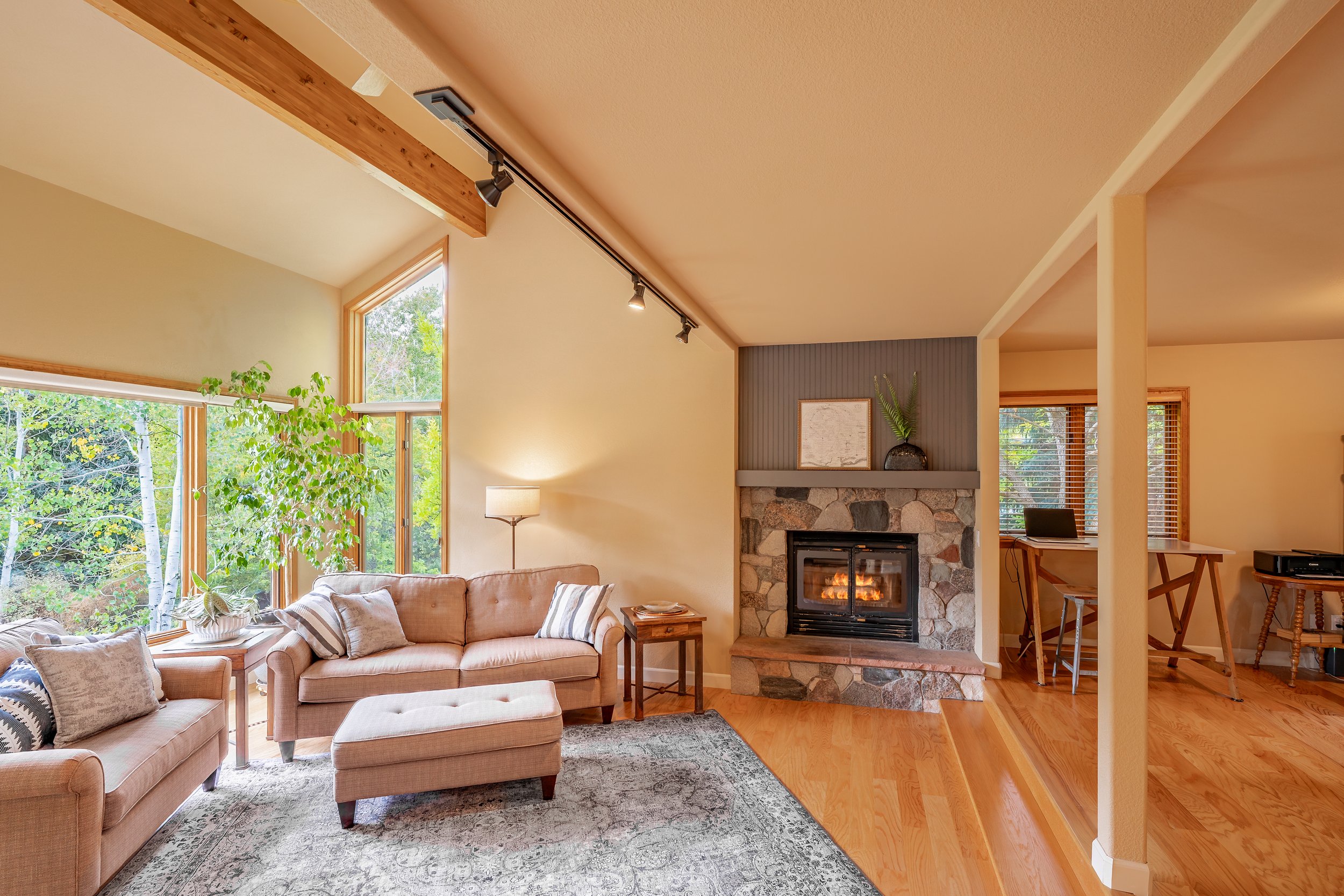 Living room with large windows, beige sofas, a stone fireplace, wooden flooring, and a small office nook with a desk and chair.