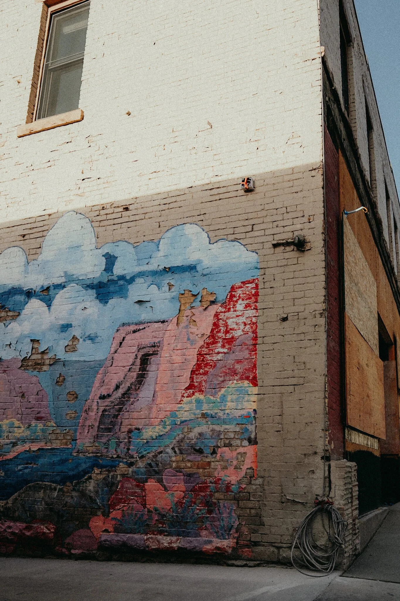 Old brick wall with a partially peeling mural of a natural landscape including clouds, a waterfall, plants, and rocks.