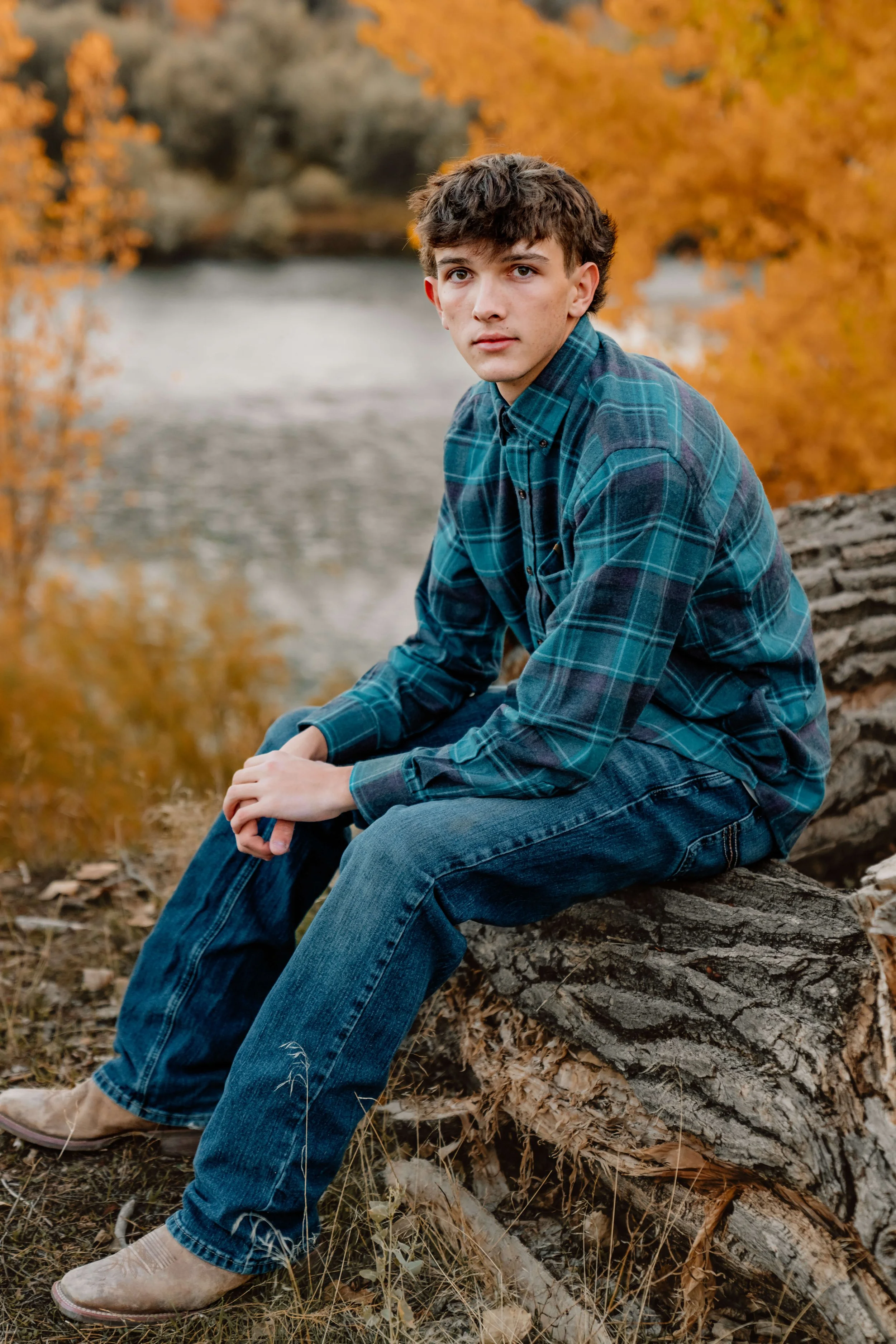 A young man with brown hair and blue eyes sitting on a fallen log by a river, surrounded by autumn foliage, wearing a blue plaid shirt and jeans.