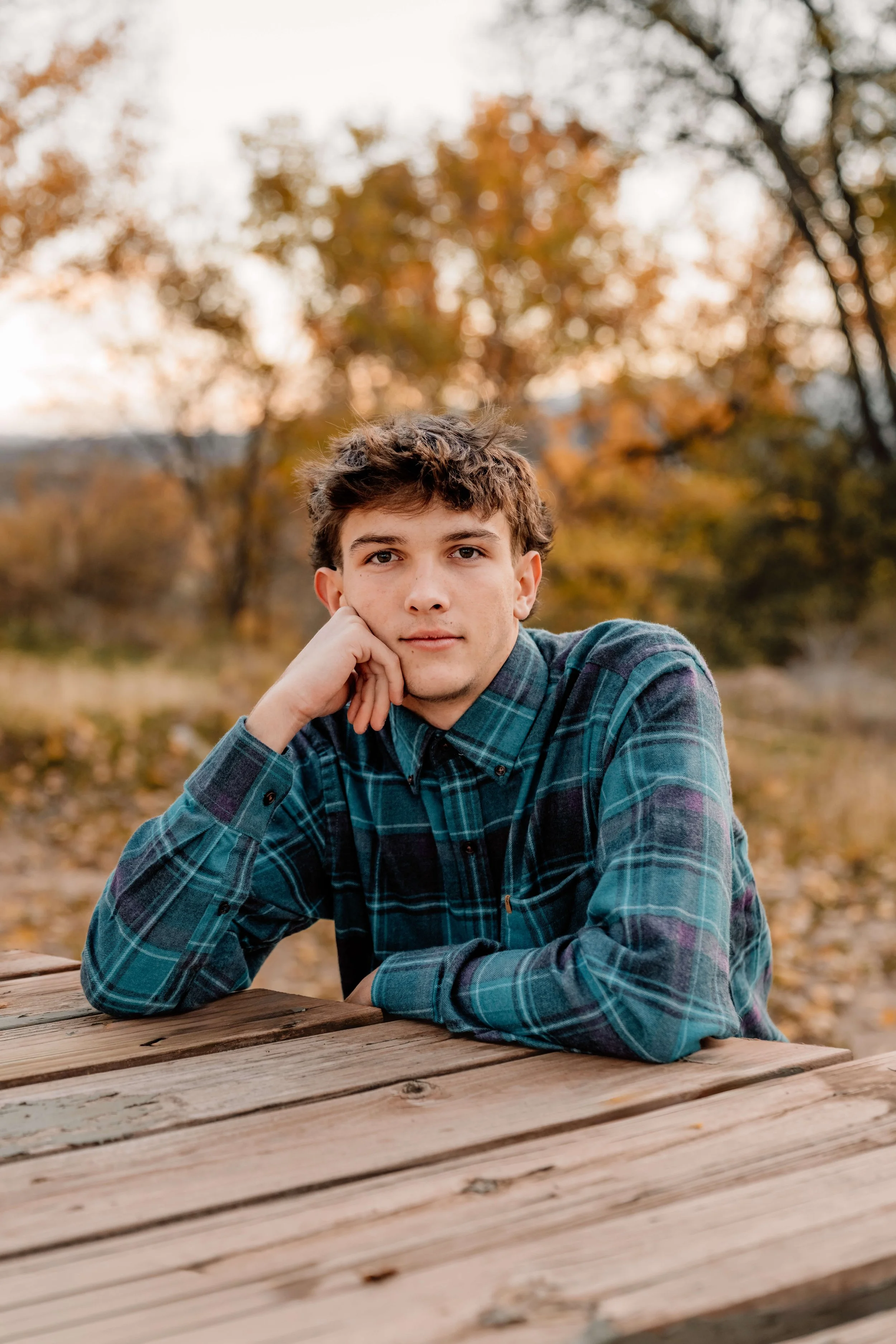 Young man in plaid shirt sitting at a wooden picnic table outdoors during fall, with autumn trees in the background