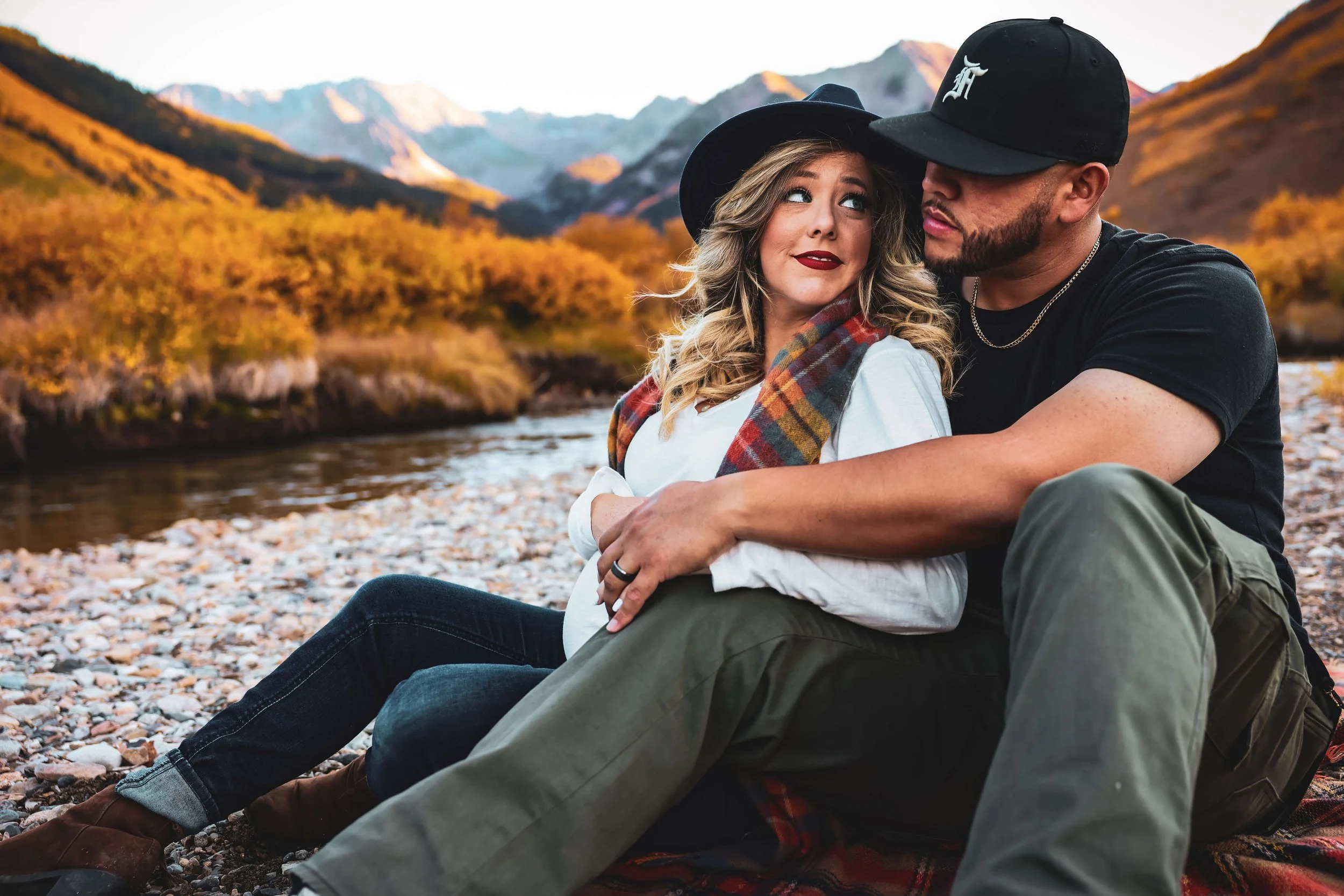 A couple sitting on a rocky riverbank surrounded by mountains and autumn trees, enjoying a moment together outdoors.