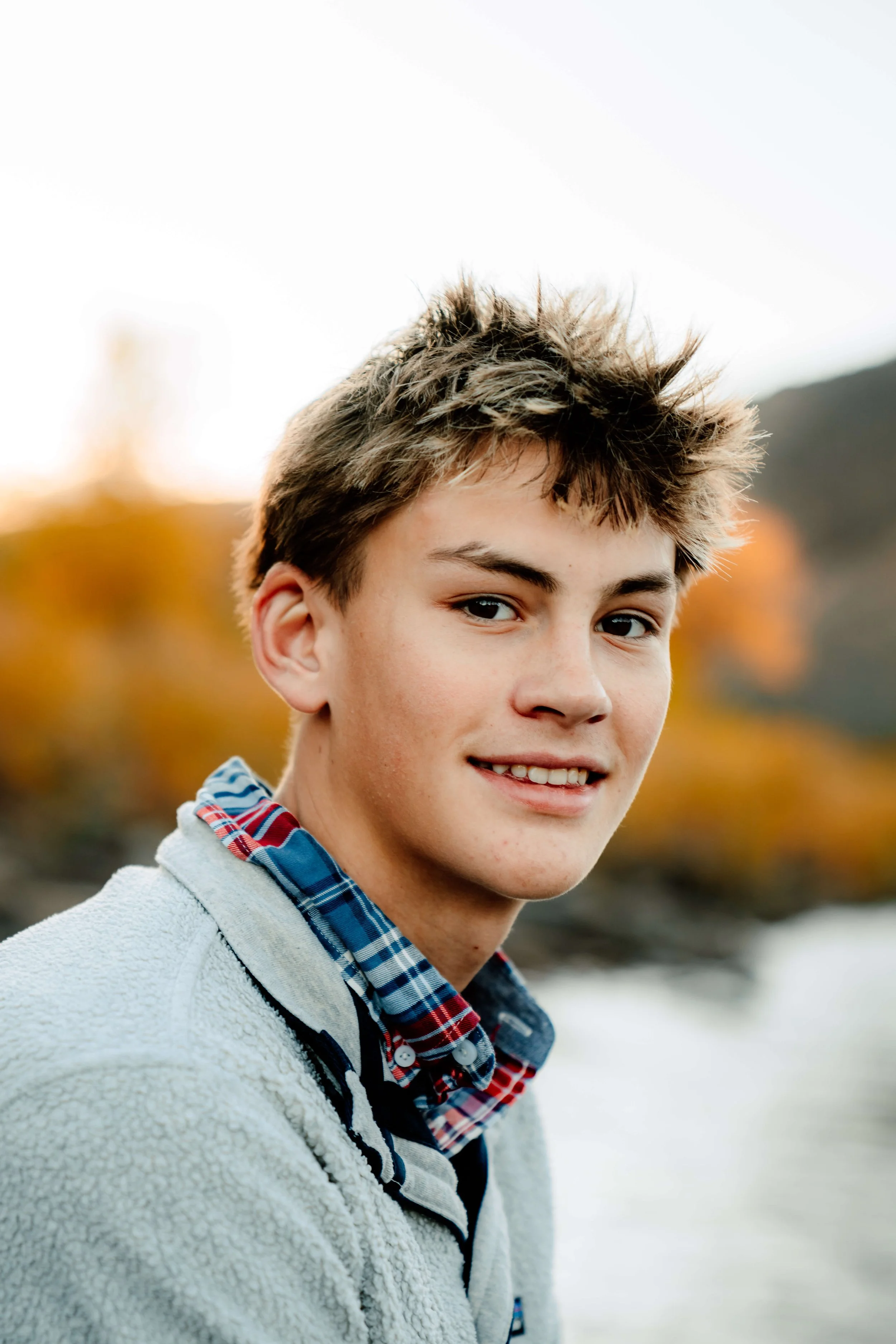 A young man with tousled brown hair, light skin, and blue eyes outdoors during autumn, wearing a plaid shirt and a light gray fleece jacket, smiling.