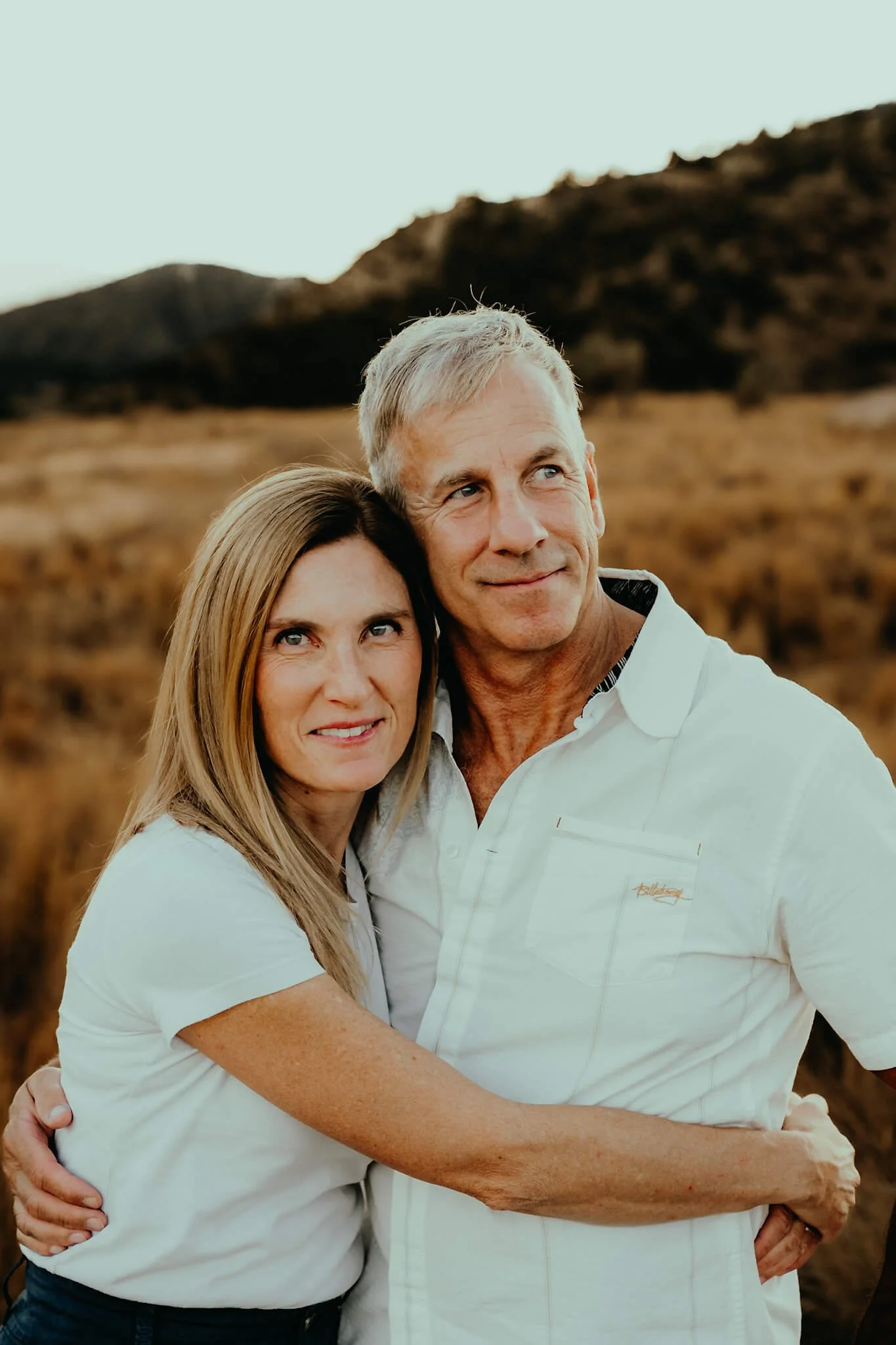 A middle-aged couple hugging outdoors in a grassy, mountainous area during sunset.