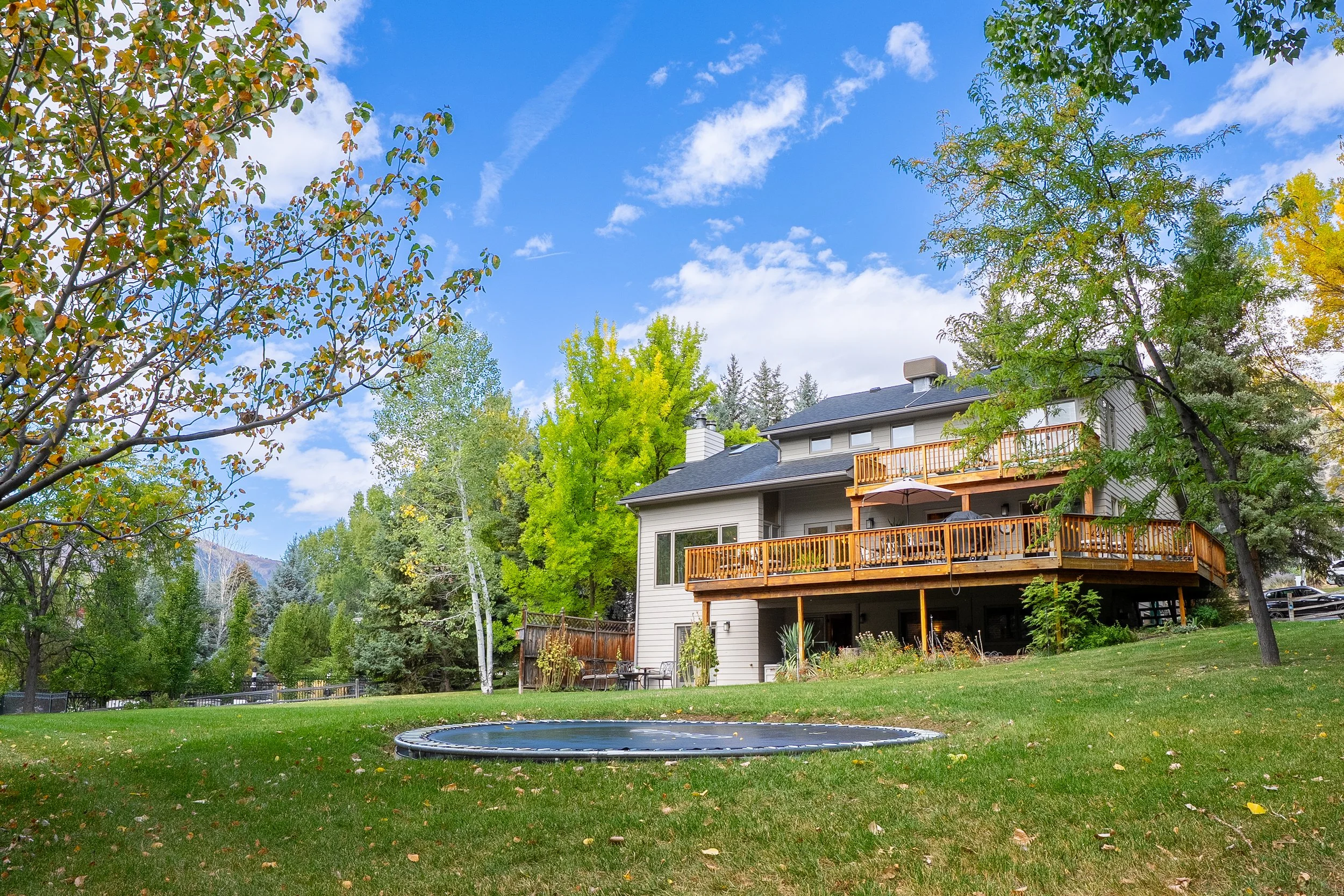A backyard with a trampoline, green grass, and trees with fall foliage in front of a two-story house with a large elevated wooden deck under a partly cloudy sky.