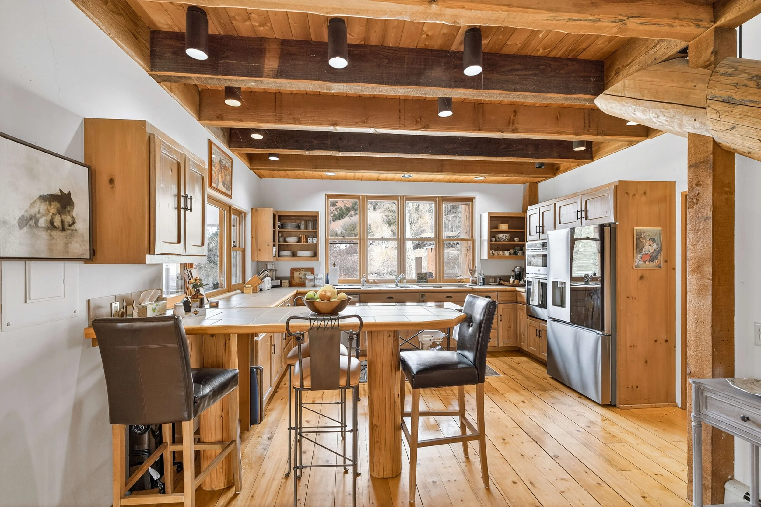 Rustic kitchen with wooden cabinets, a central island with bar stools, kitchen appliances, and large windows letting in natural light.