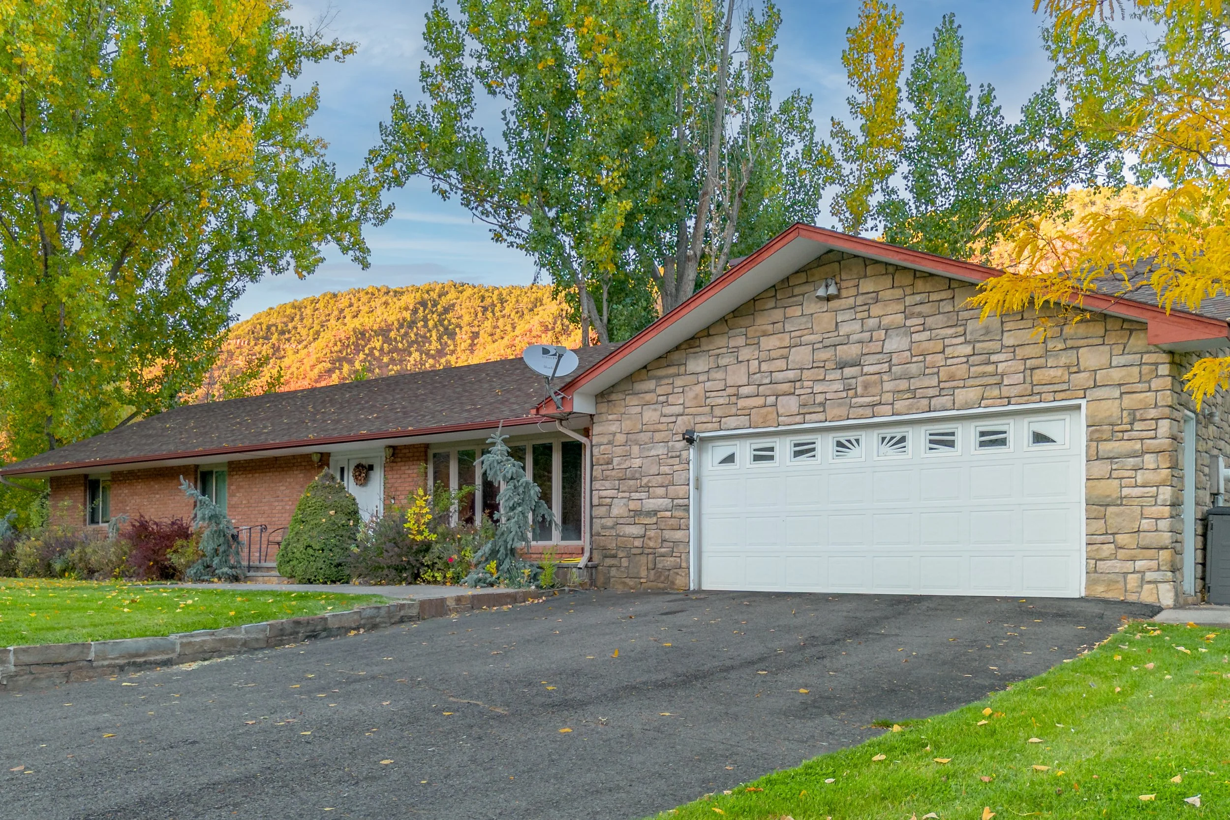 A suburban house with a brick exterior, stone accents, a white garage door, and a driveway, surrounded by green trees and a lawn, with hills and a partly cloudy sky in the background.