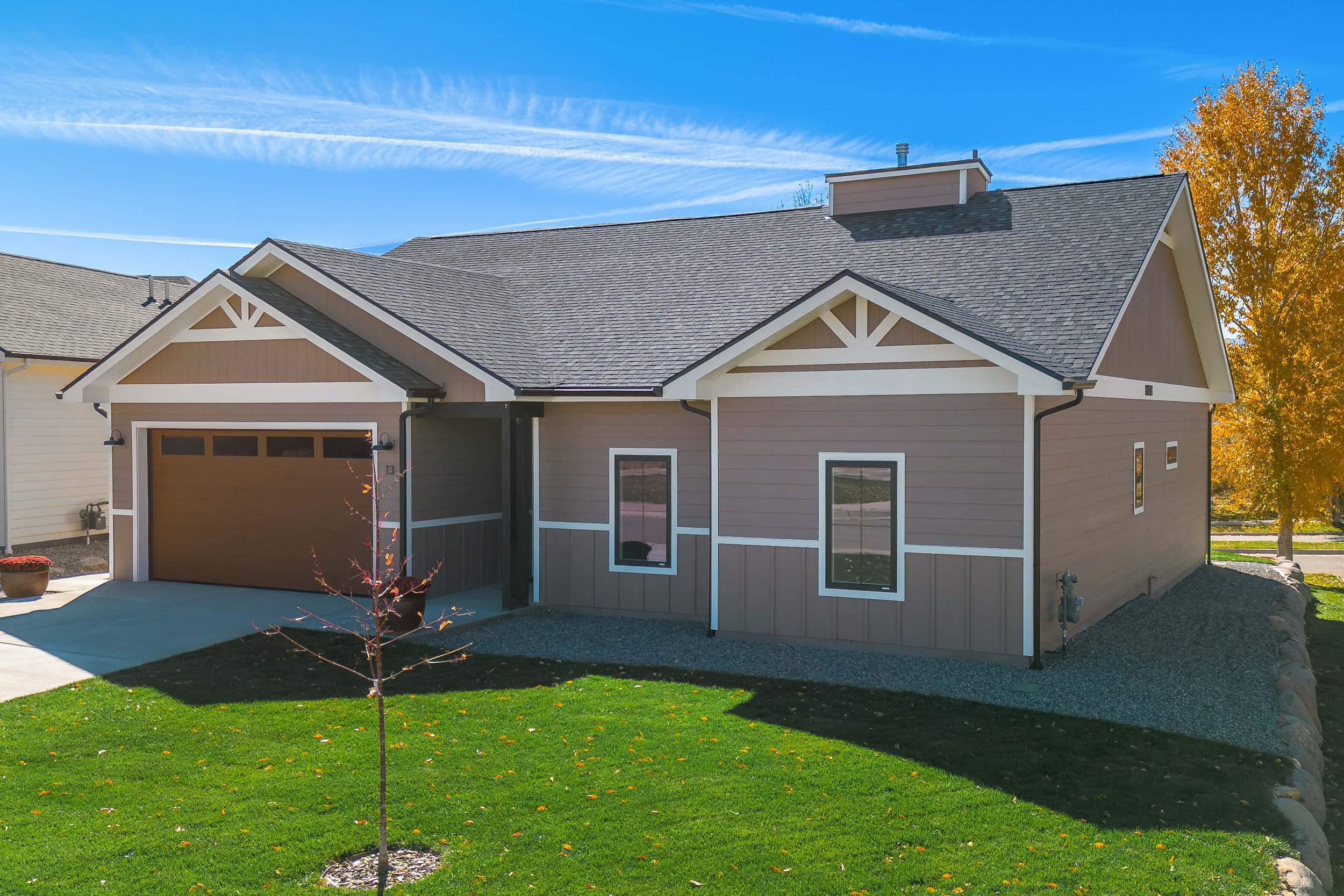 A modern house with brown and beige exterior, a garage door, and a small front yard with green grass and a young tree, under a blue sky with clouds and trees with autumn leaves in the background.