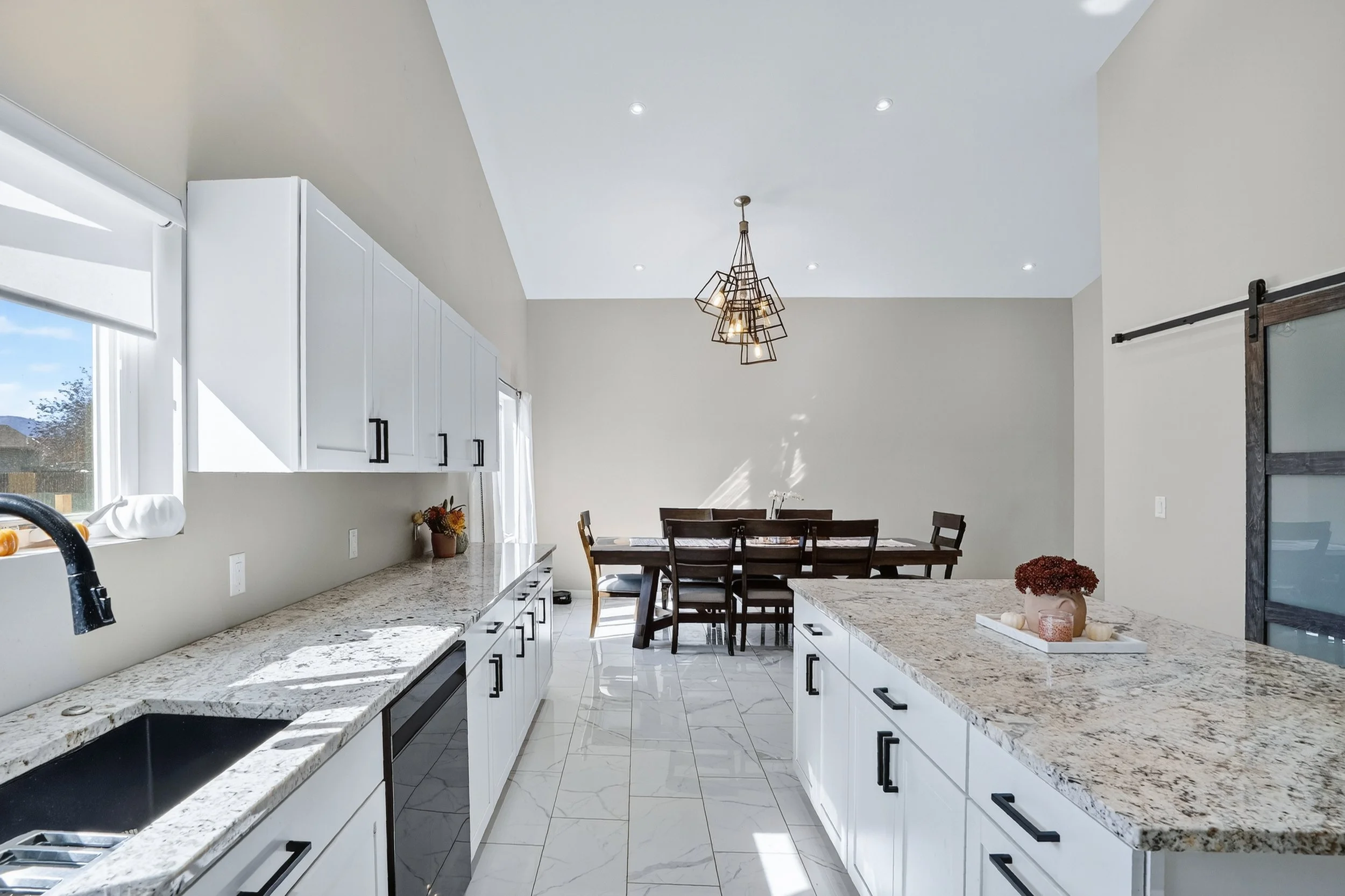 Modern kitchen with white cabinets, granite countertops, and a dining area with a wooden table and chairs. Pendant light fixture hangs above the table. Window on the left side allowing natural light.
