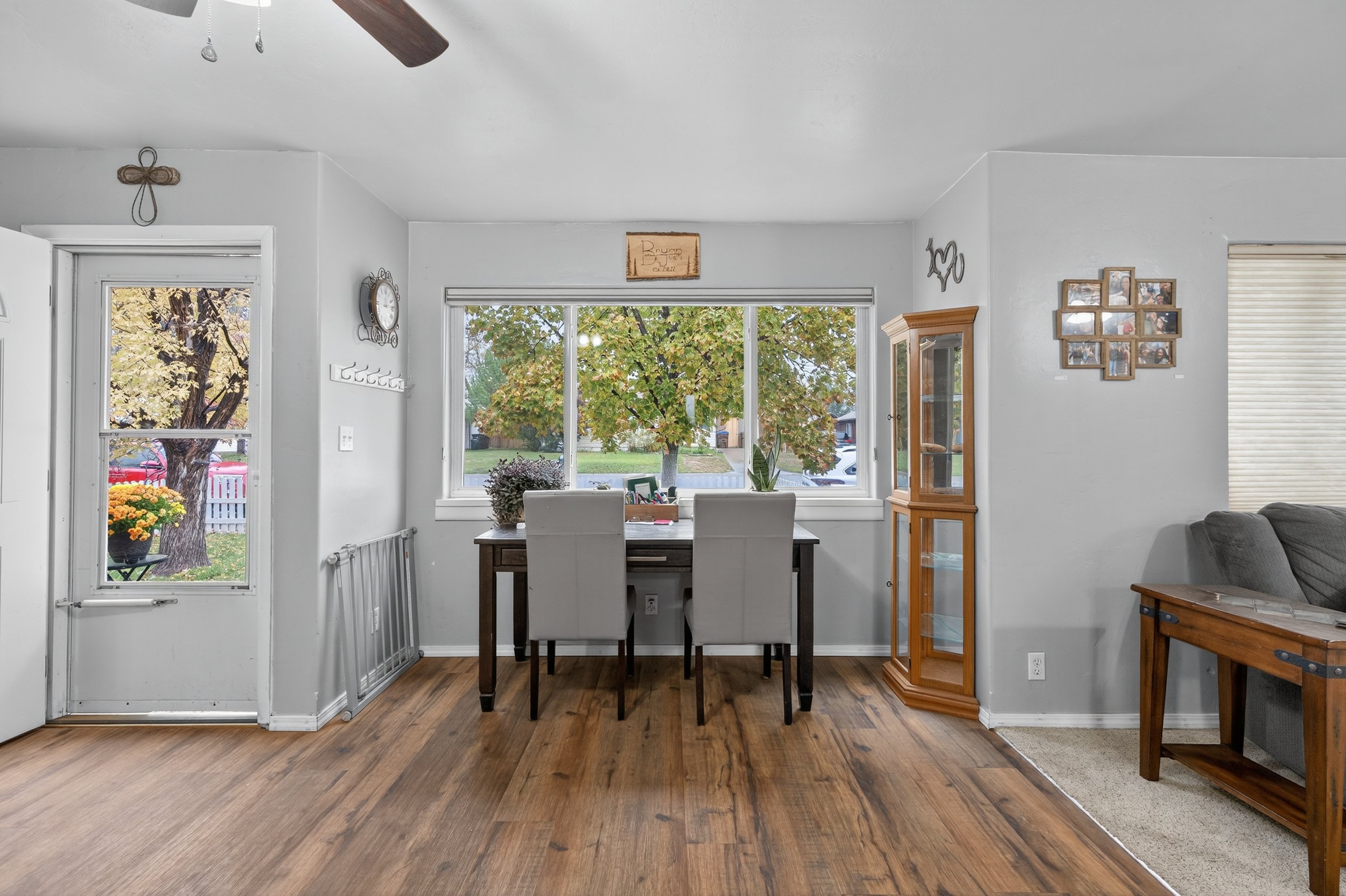 Living room with a wooden floor, a dining table with four white chairs, large window showing an outdoor tree, a wooden corner cabinet, framed photos on the wall, and part of a gray sofa on the right.